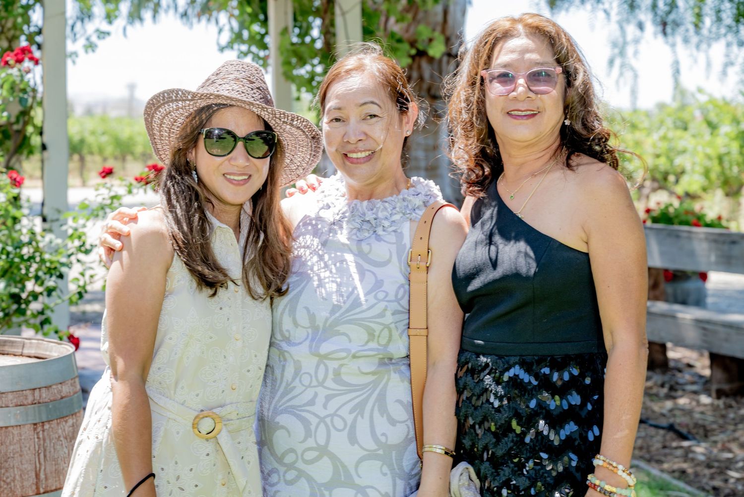 Three women smiling together outdoors at a sunny garden event, one wearing a straw hat and sunglasses.