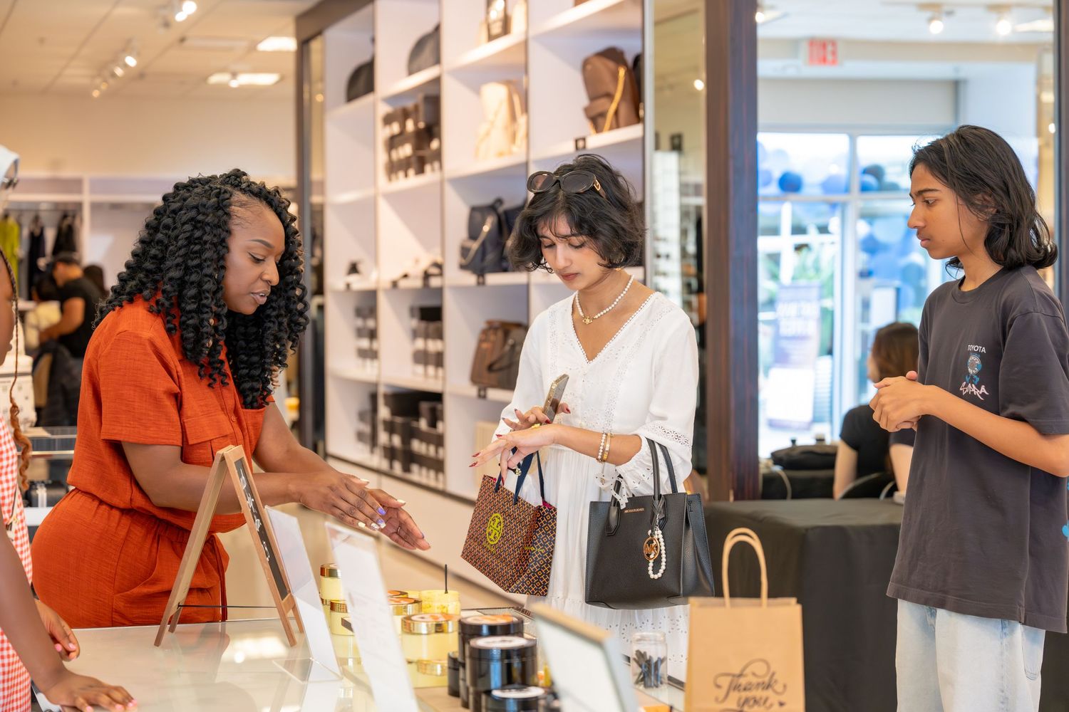 A store associate assists two customers at a retail shop counter, with shelves of products displayed in the background.