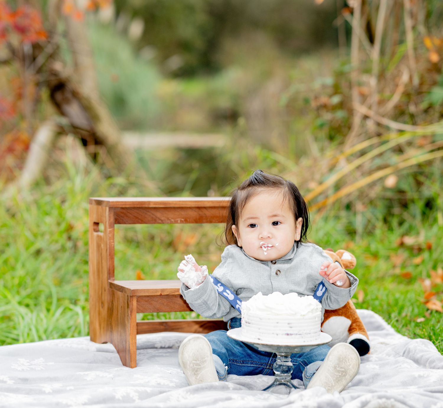 Messy baby boy enjoys first birthday smash cake outdoors on white blanket in autumn setting.