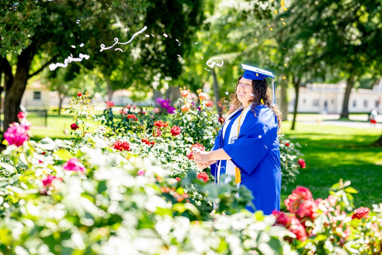 Smiling female graduate in blue cap and gown posing among vibrant roses in sunny park.