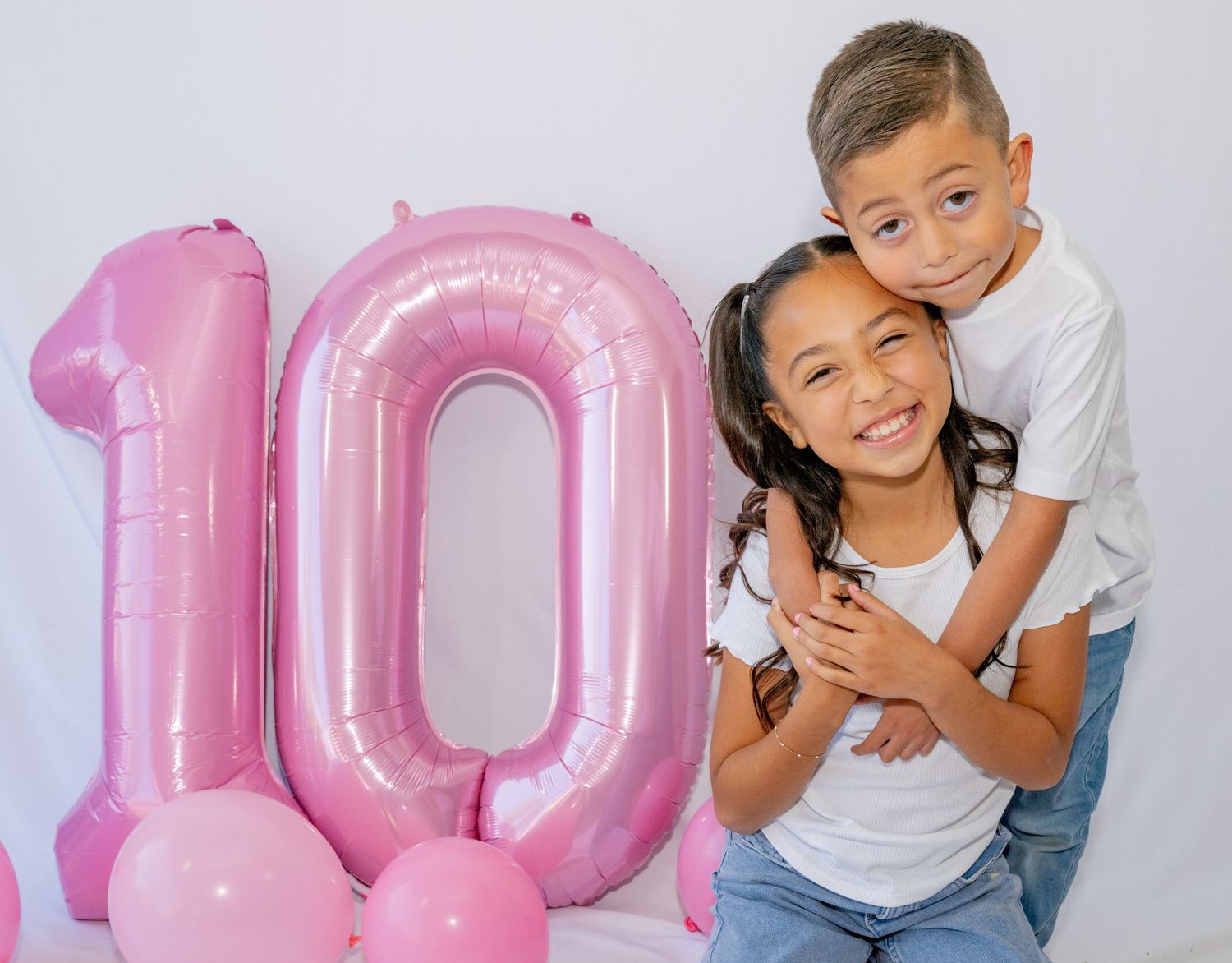 Boy hugs smiling girl from behind beside large pink number 10 birthday balloon.