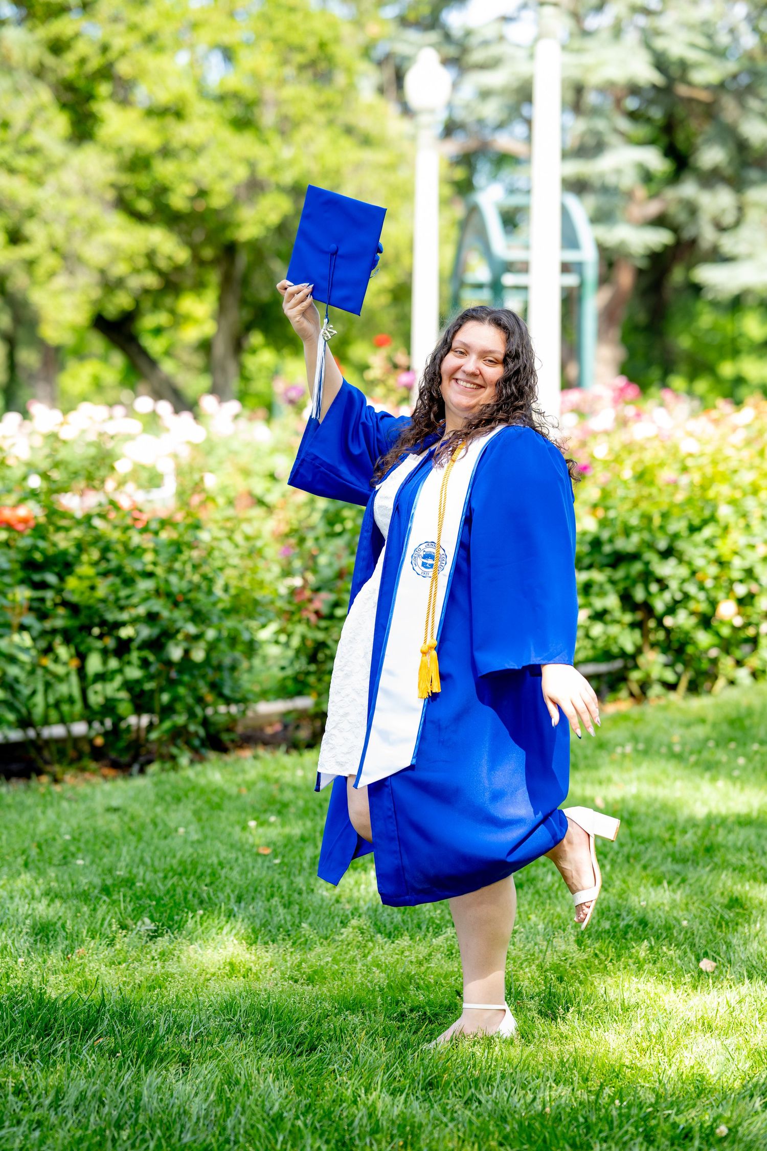 Joyful graduate in blue cap and gown tosses cap while celebrating outdoors in a garden.