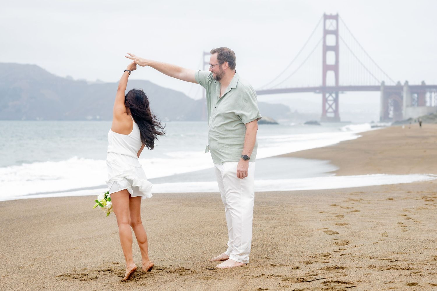 Romantic couple twirling on San Francisco Baker Beach with iconic Golden Gate Bridge in background.