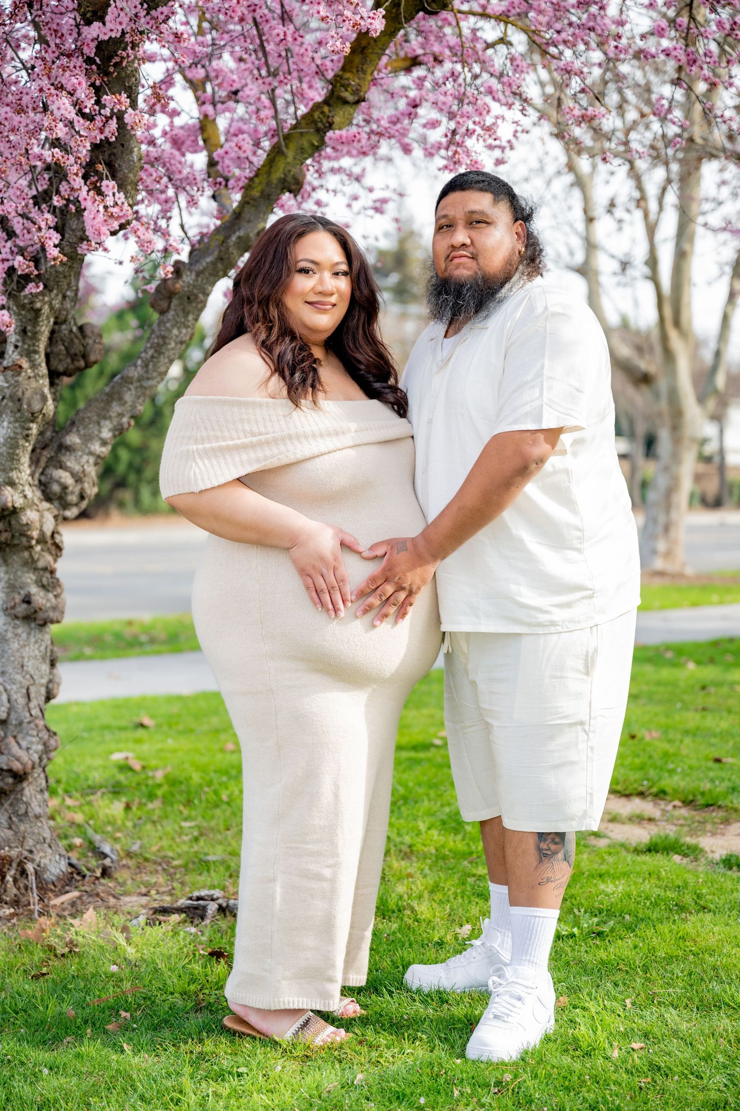 Maternity photo of couple in white outfits holding baby bump under pink spring blossoms.