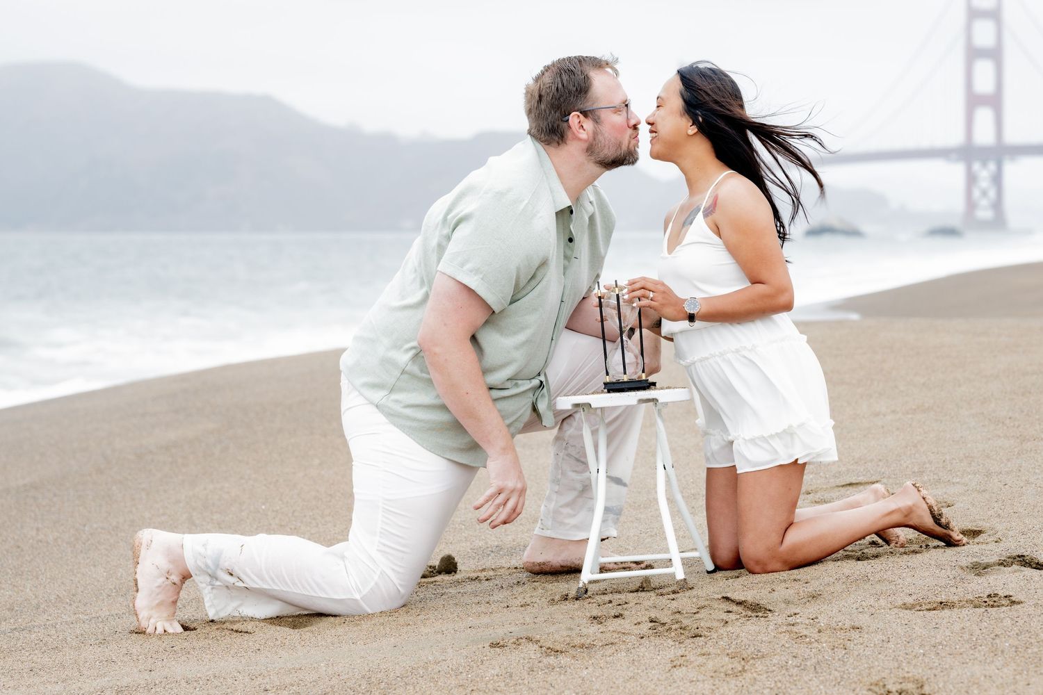 Couple sharing a kiss while kneeling on Baker Beach with Golden Gate Bridge in the background.