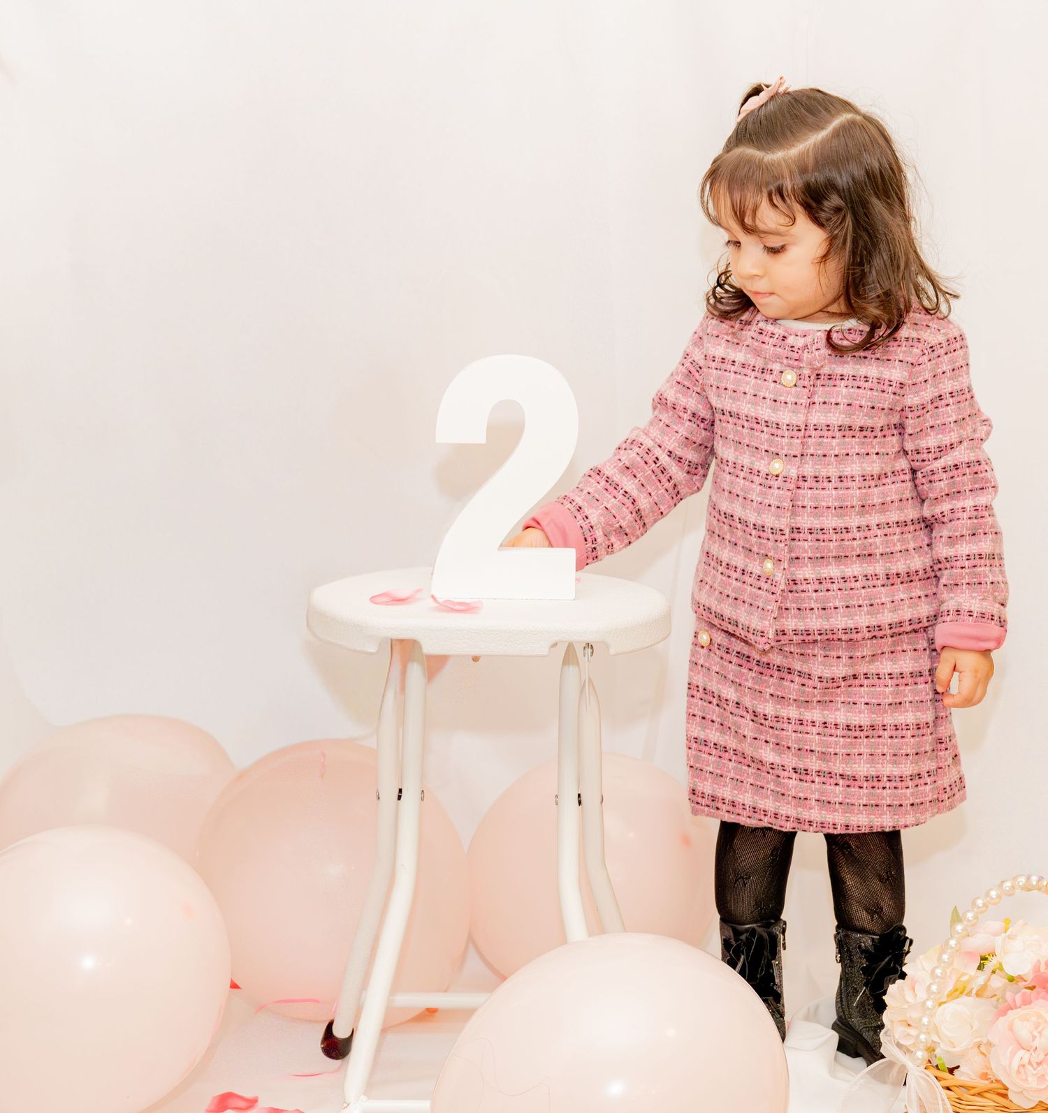 Toddler girl in pink tweed dress stands by white table with number 2, surrounded by pink balloons at birthday party.