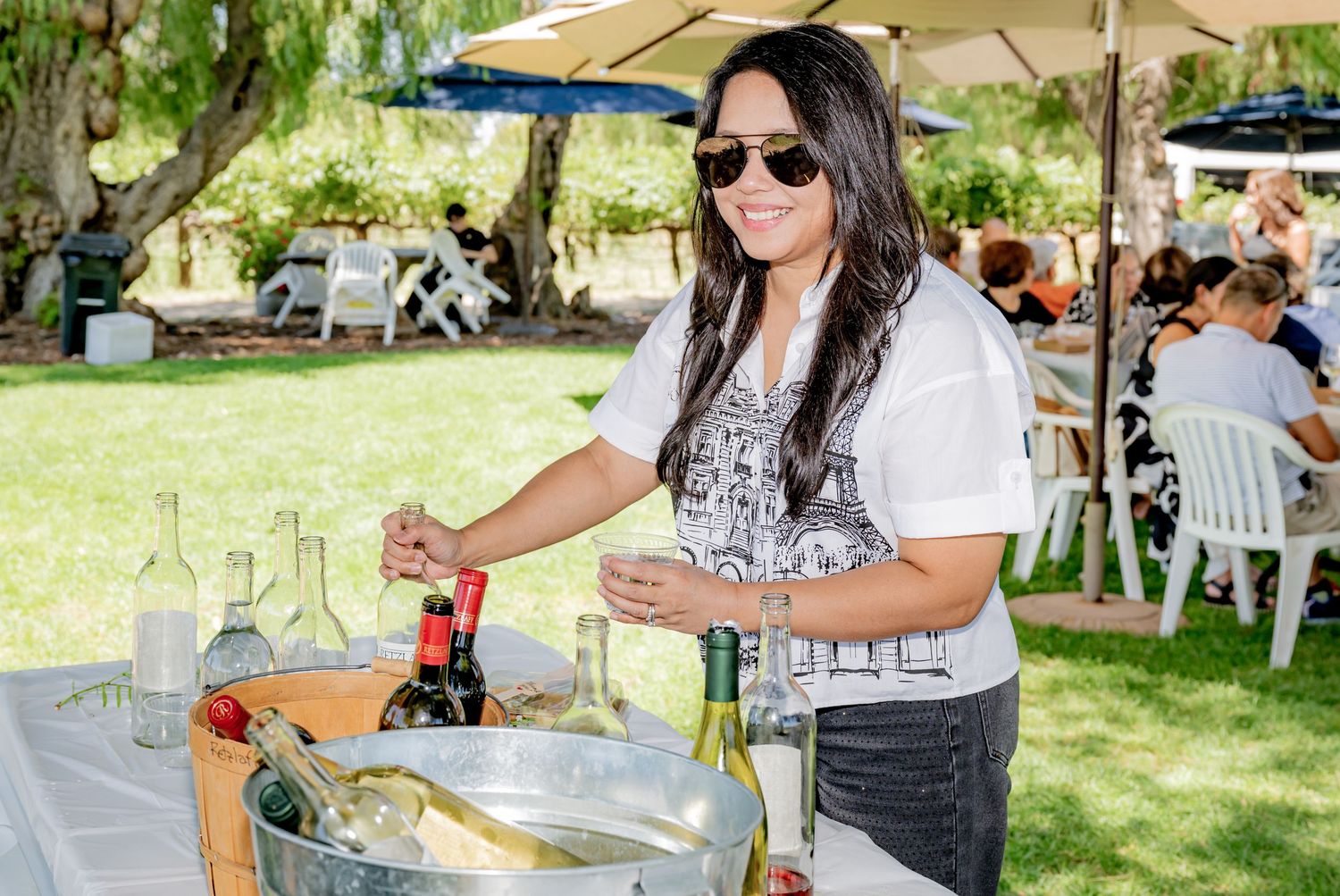 Woman in sunglasses serving wine at an outdoor event with bottles chilling in an ice bucket.