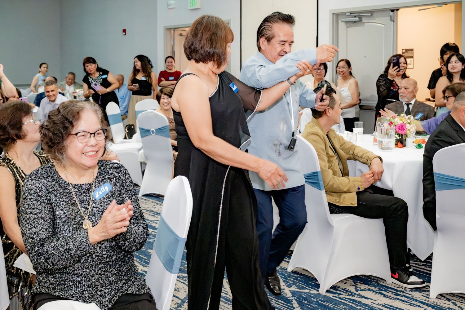 People dancing and socializing at indoor banquet event with guests seated at white chair-covered tables.
