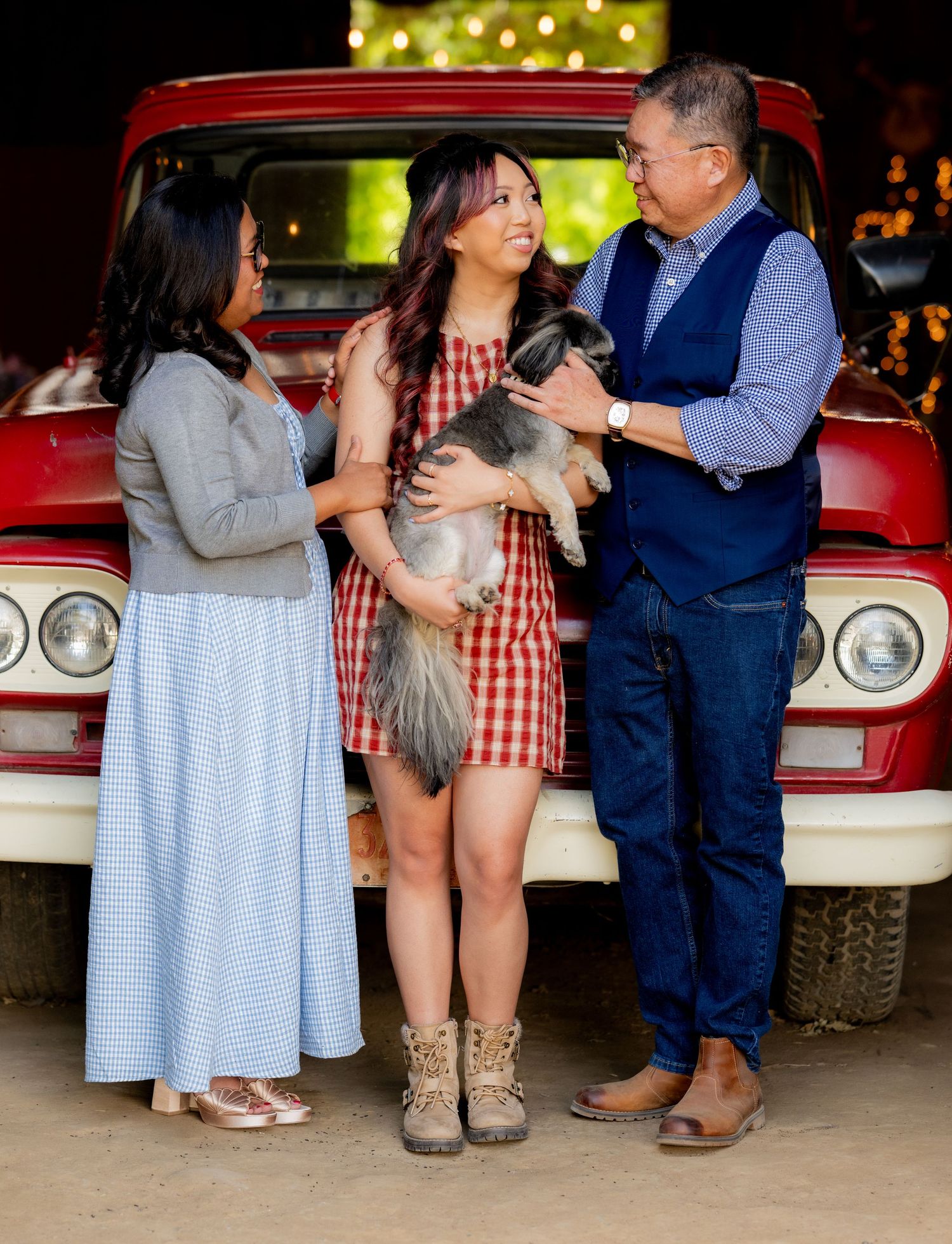 Family of three with small dog laughing together in front of vintage red truck indoors.