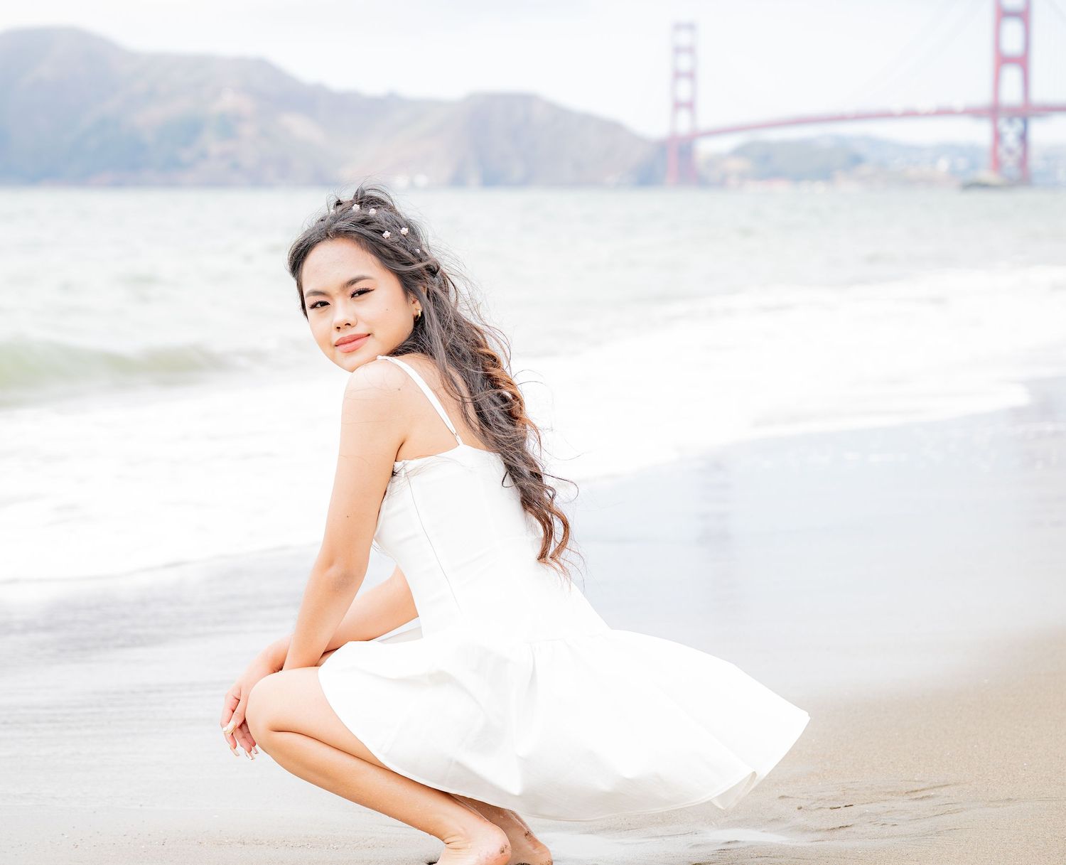 Young woman in white dress crouching on Baker Beach shoreline, smiling, with Golden Gate Bridge behind her.