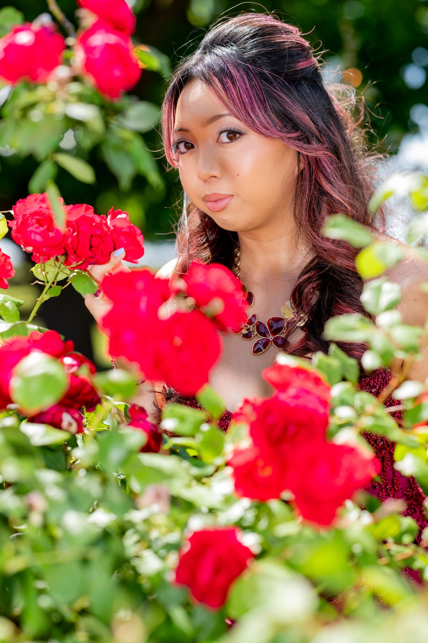 Young Asian woman with highlights gazes at vibrant red roses in a lush garden.