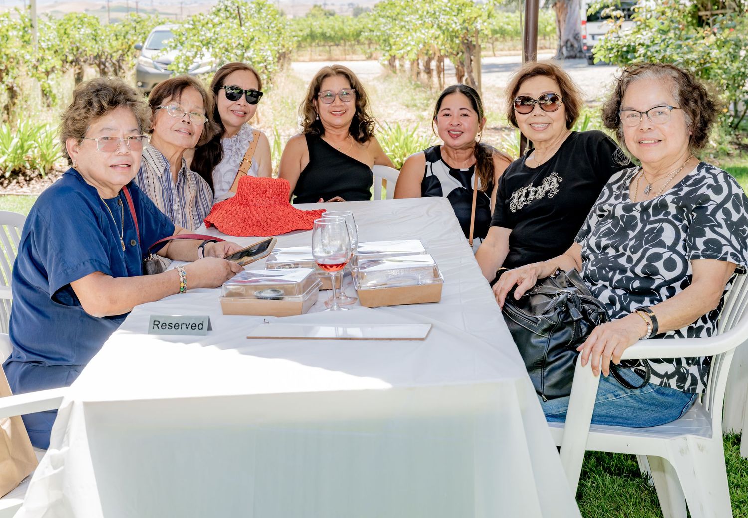 Group of women smiling together at a reserved outdoor table with drinks on a sunny day.