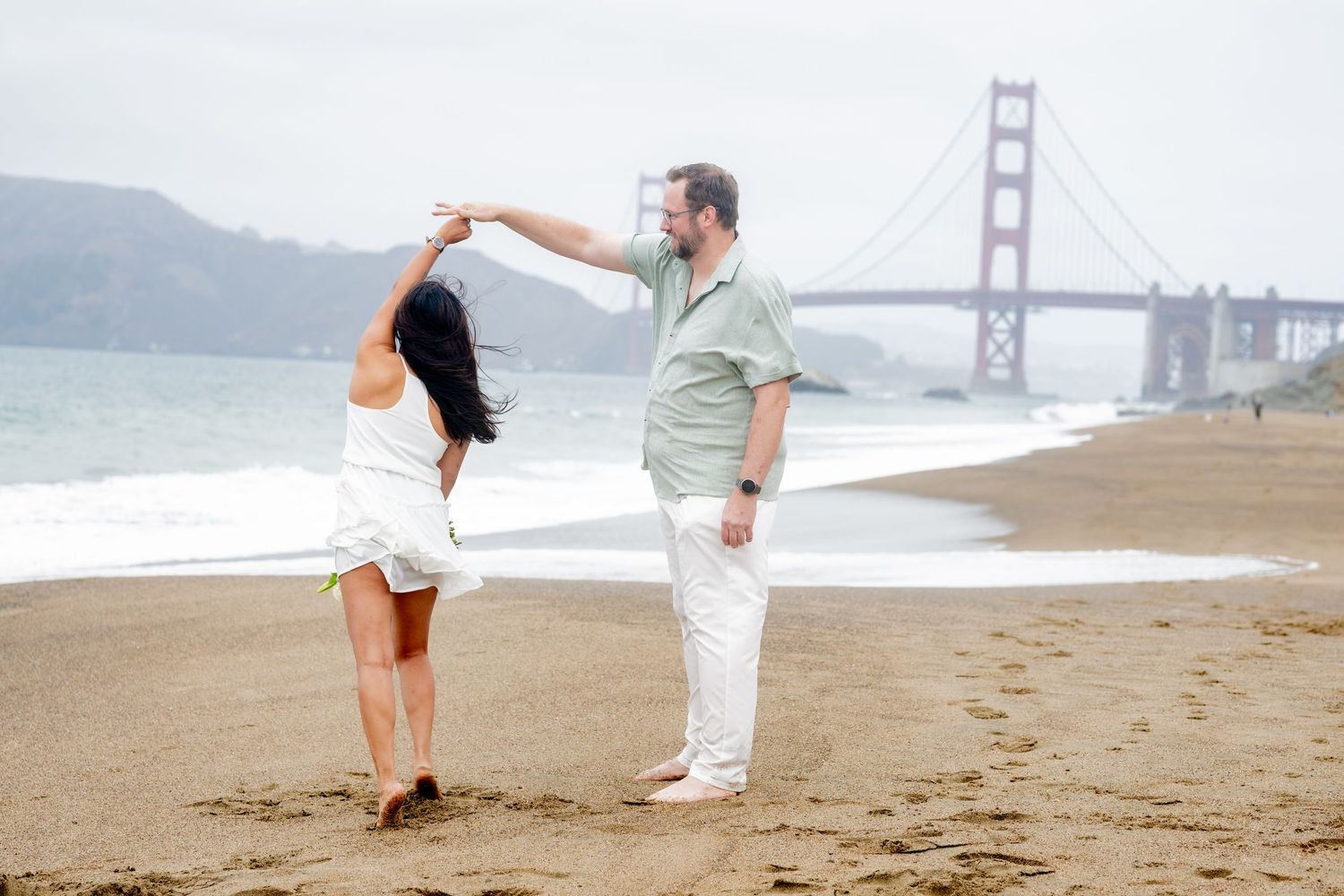 Joyful couple dancing barefoot on Baker Beach with the Golden Gate Bridge in the misty background.
