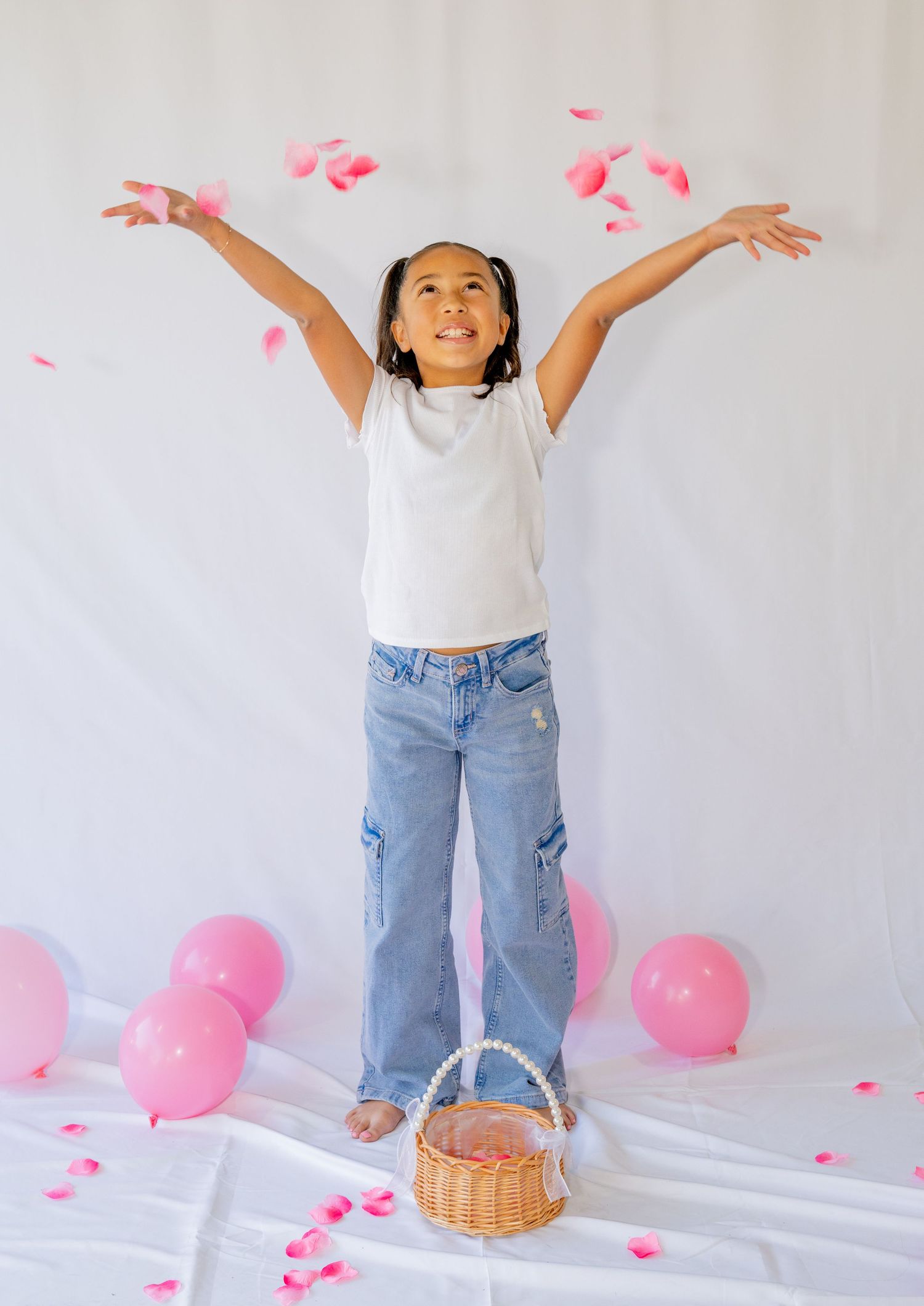 Joyful girl throwing pink rose petals in air with arms raised surrounded by pink balloons indoors.
