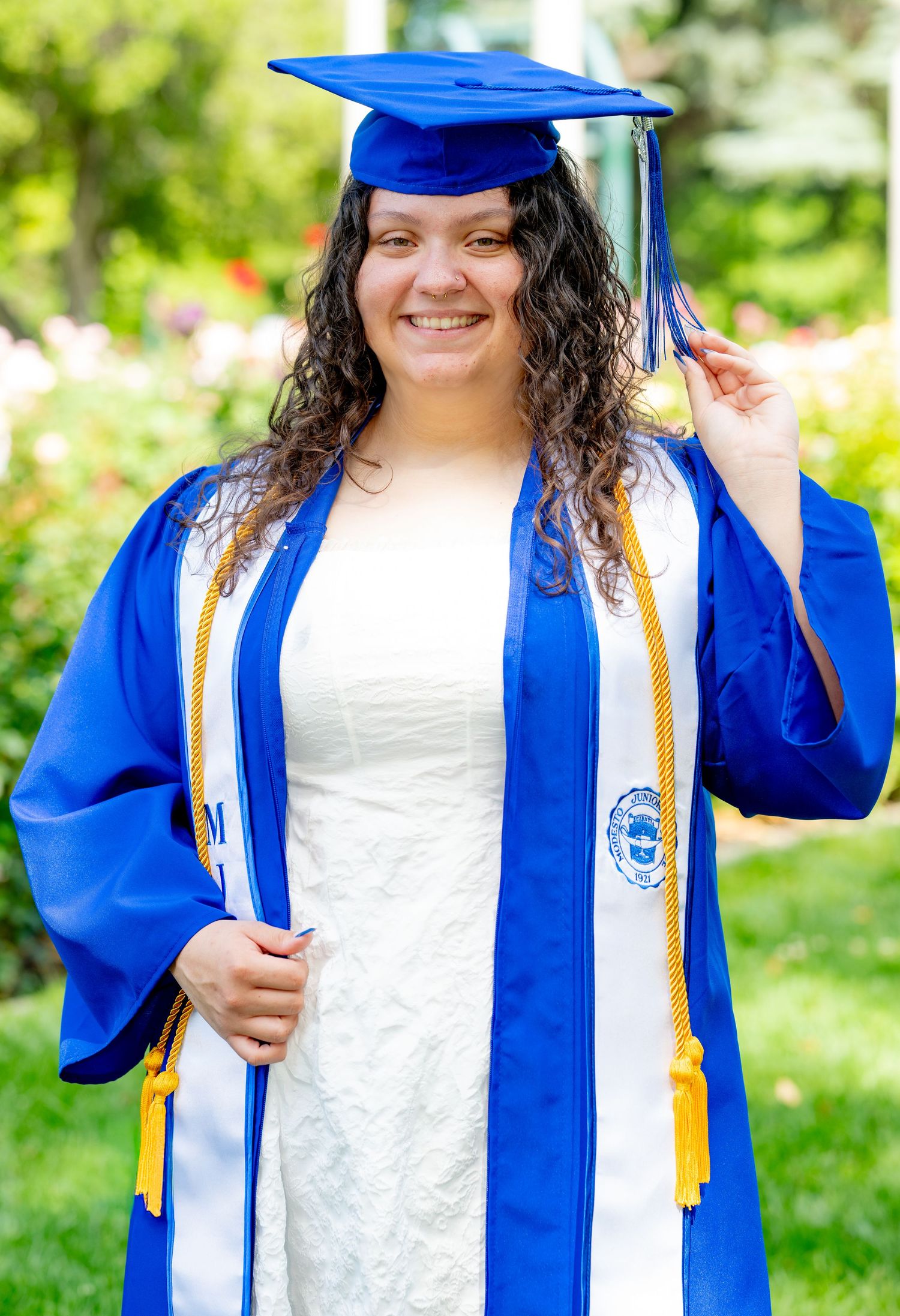 Smiling graduate poses in blue cap and gown with gold honor cords in a garden setting.