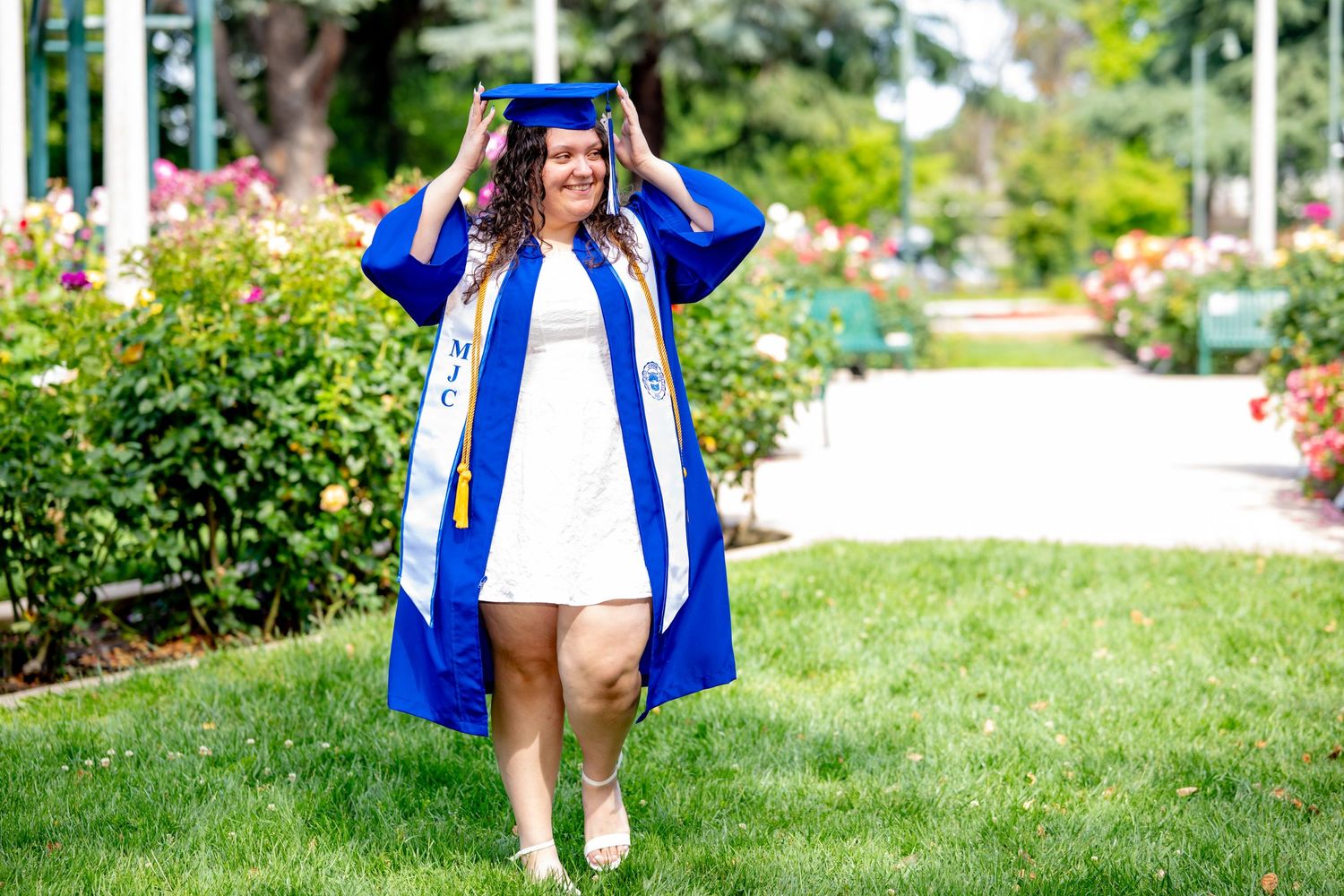 Smiling graduate adjusts her blue cap while walking through a colorful rose garden.