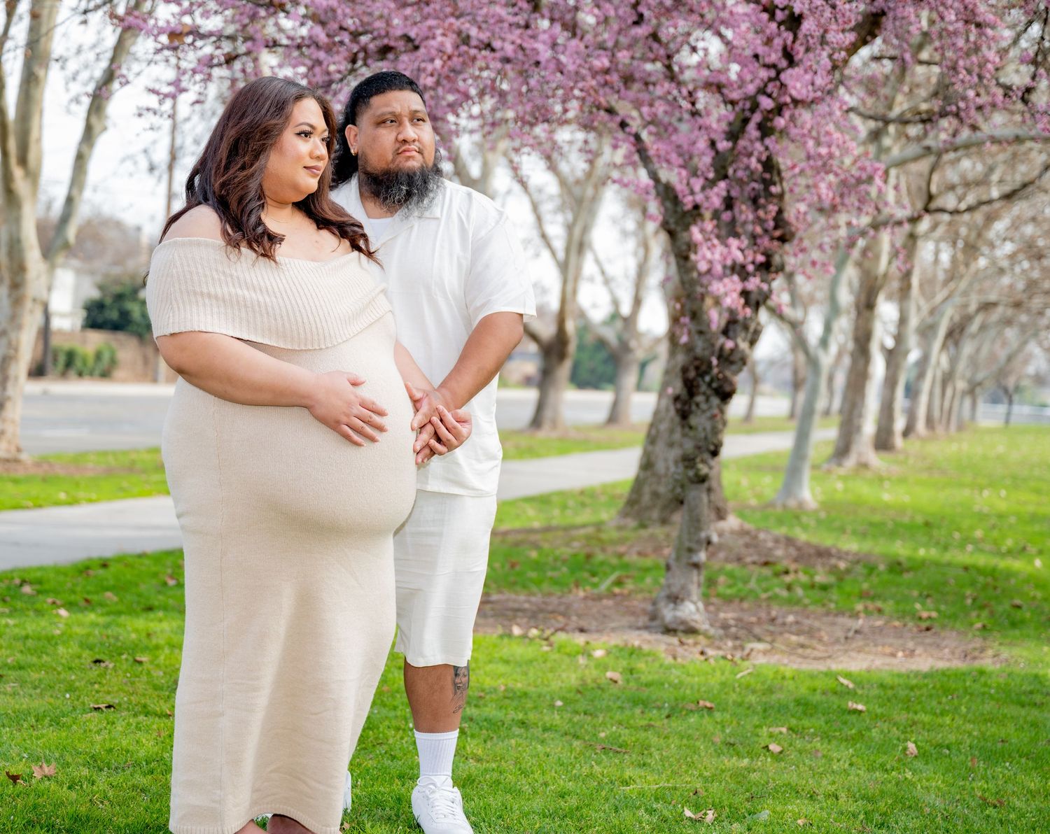 Pregnant woman in beige knit dress with partner standing under pink flowering trees in park.