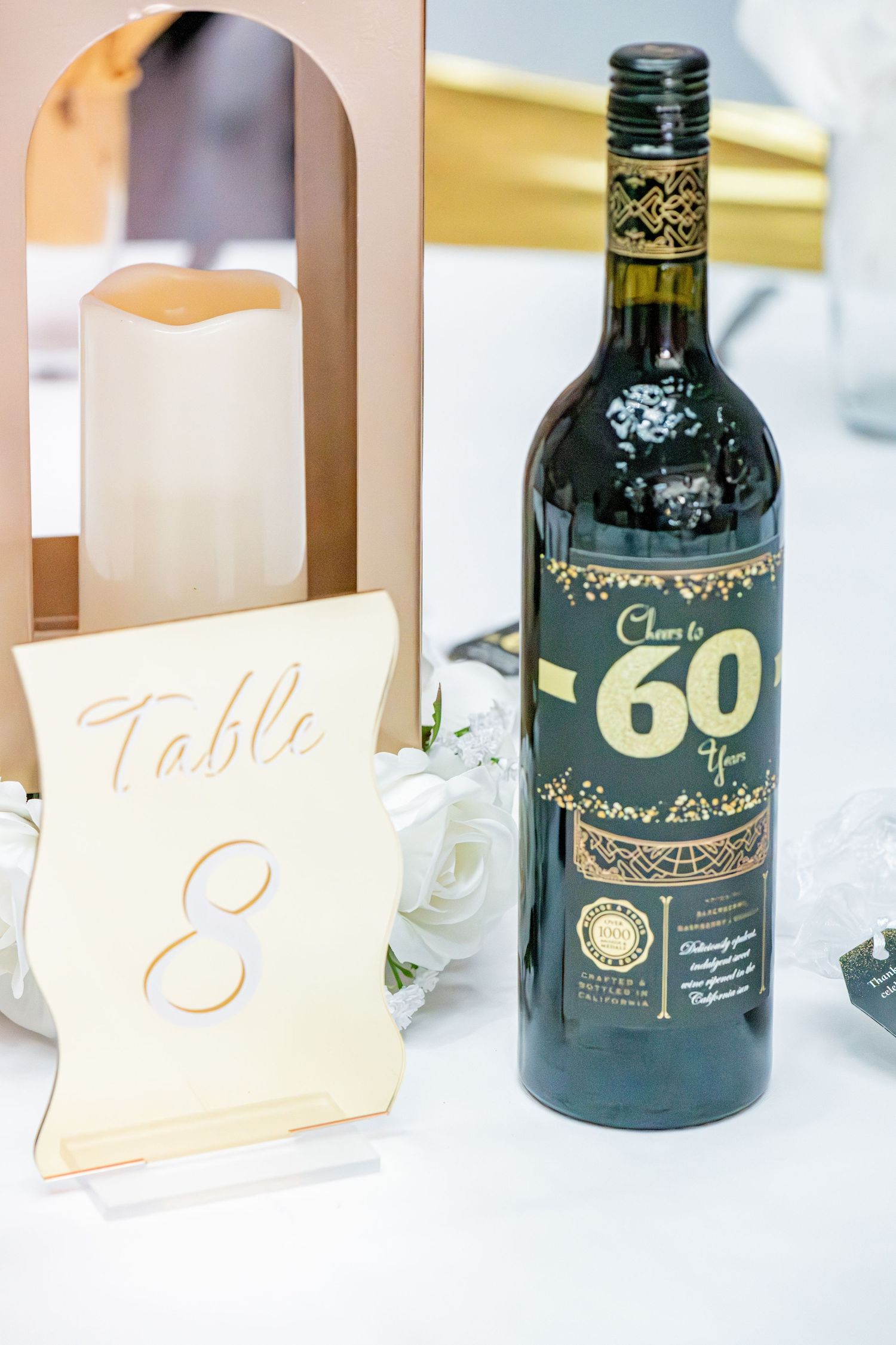 Elegant 60th birthday party table setting with wine bottle, white roses, and table number eight.