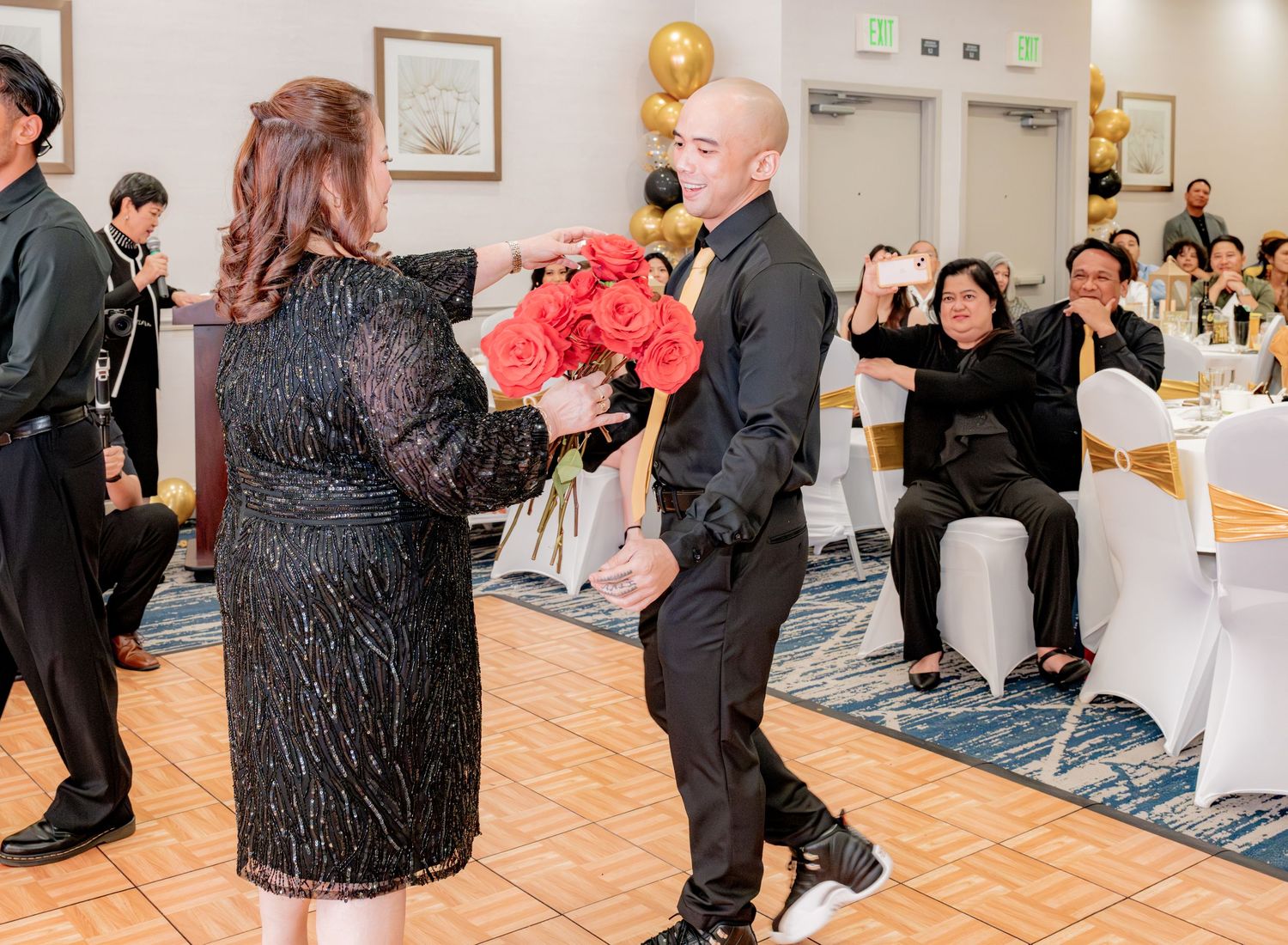 Man in black suit presents bouquet of red roses to woman at formal 60th birthday party celebration.