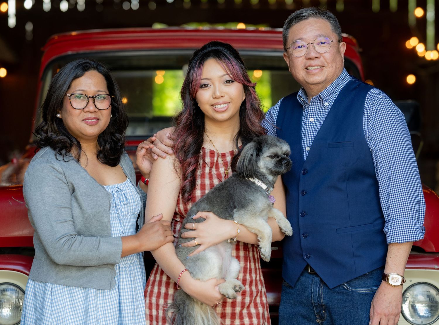 Family of three smiling with small gray dog posed in front of classic red pickup truck.
