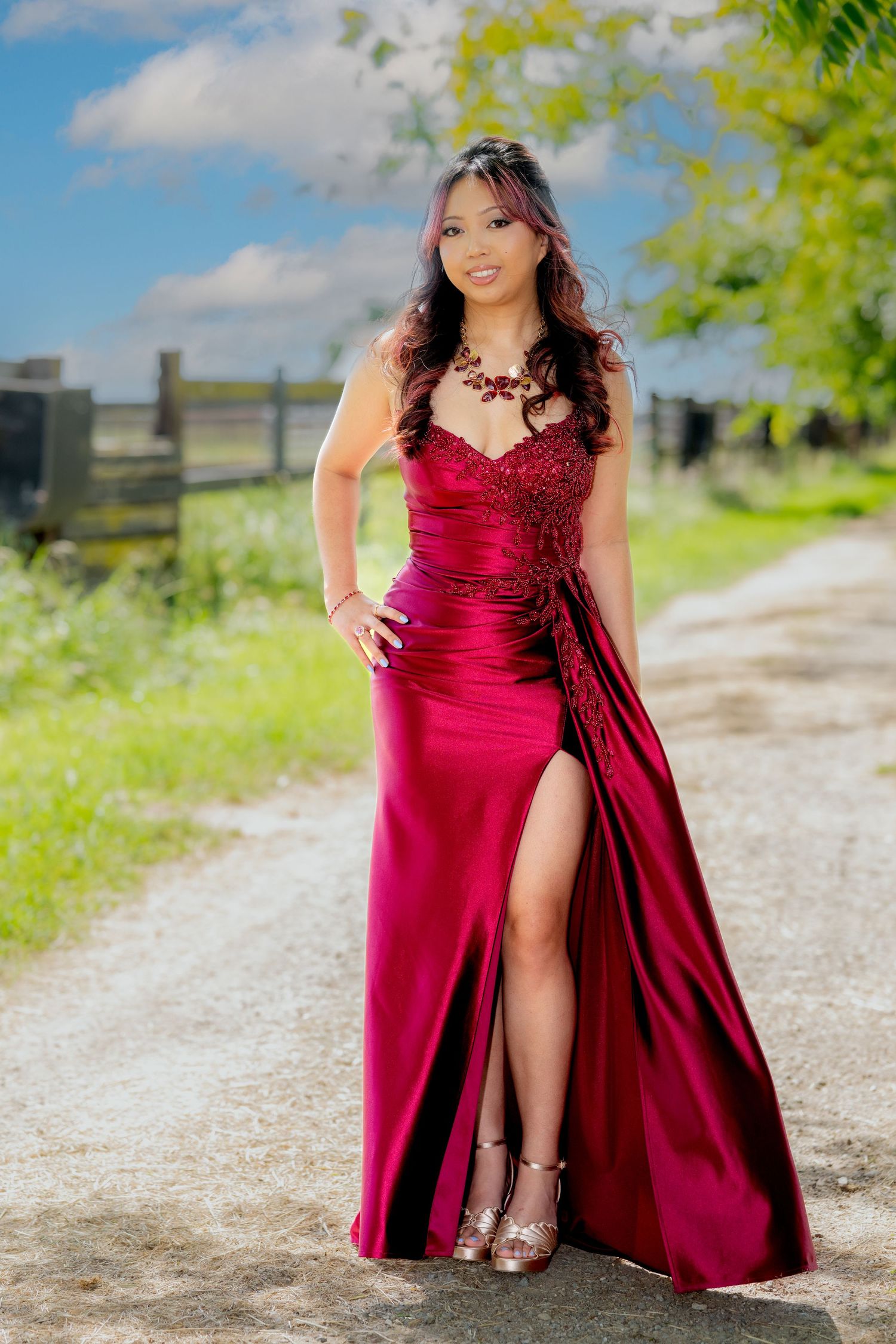 Young woman in elegant red satin gown with slit poses on a country dirt road.