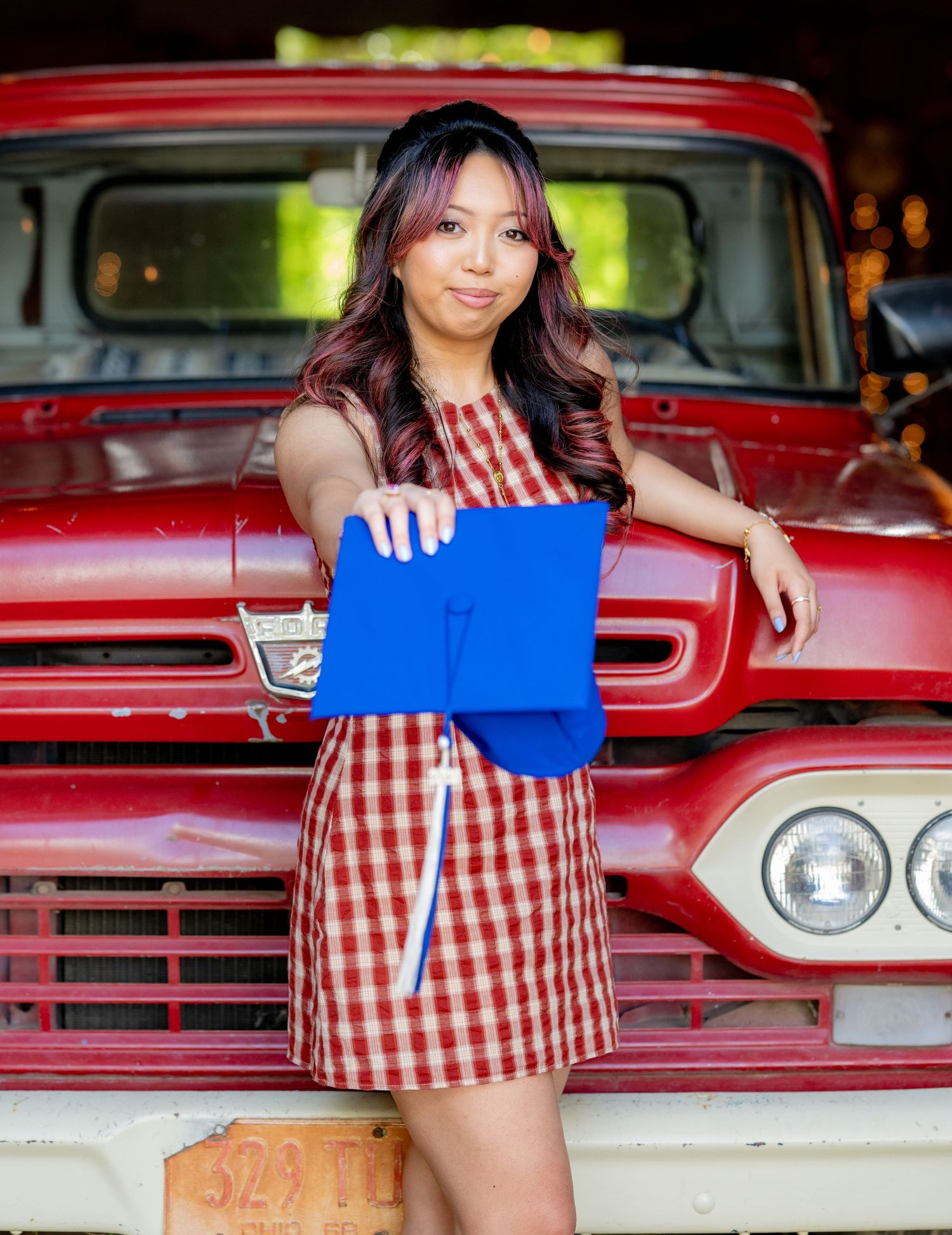 Smiling graduate in plaid dress leans on classic red truck holding blue cap and diploma.