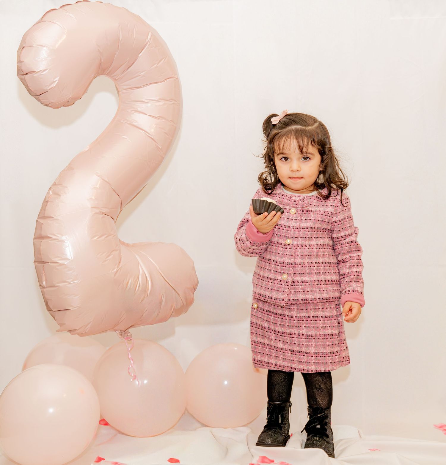 A toddler girl in a pink tweed dress stands beside a large number 2 balloon at her 2nd birthday party.