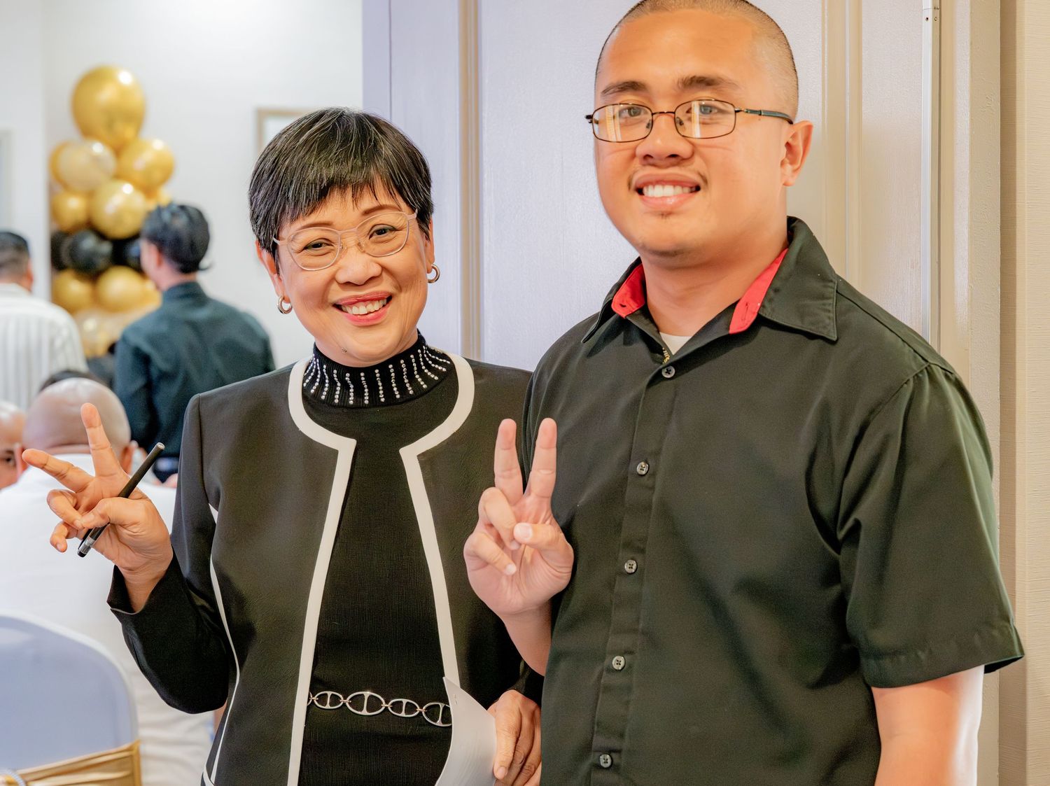 Two people smiling and making peace signs at an indoor party.