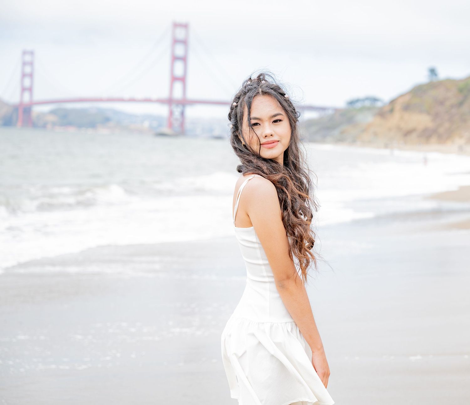 Young woman in white dress posing on Baker Beach with iconic Golden Gate Bridge visible behind her.