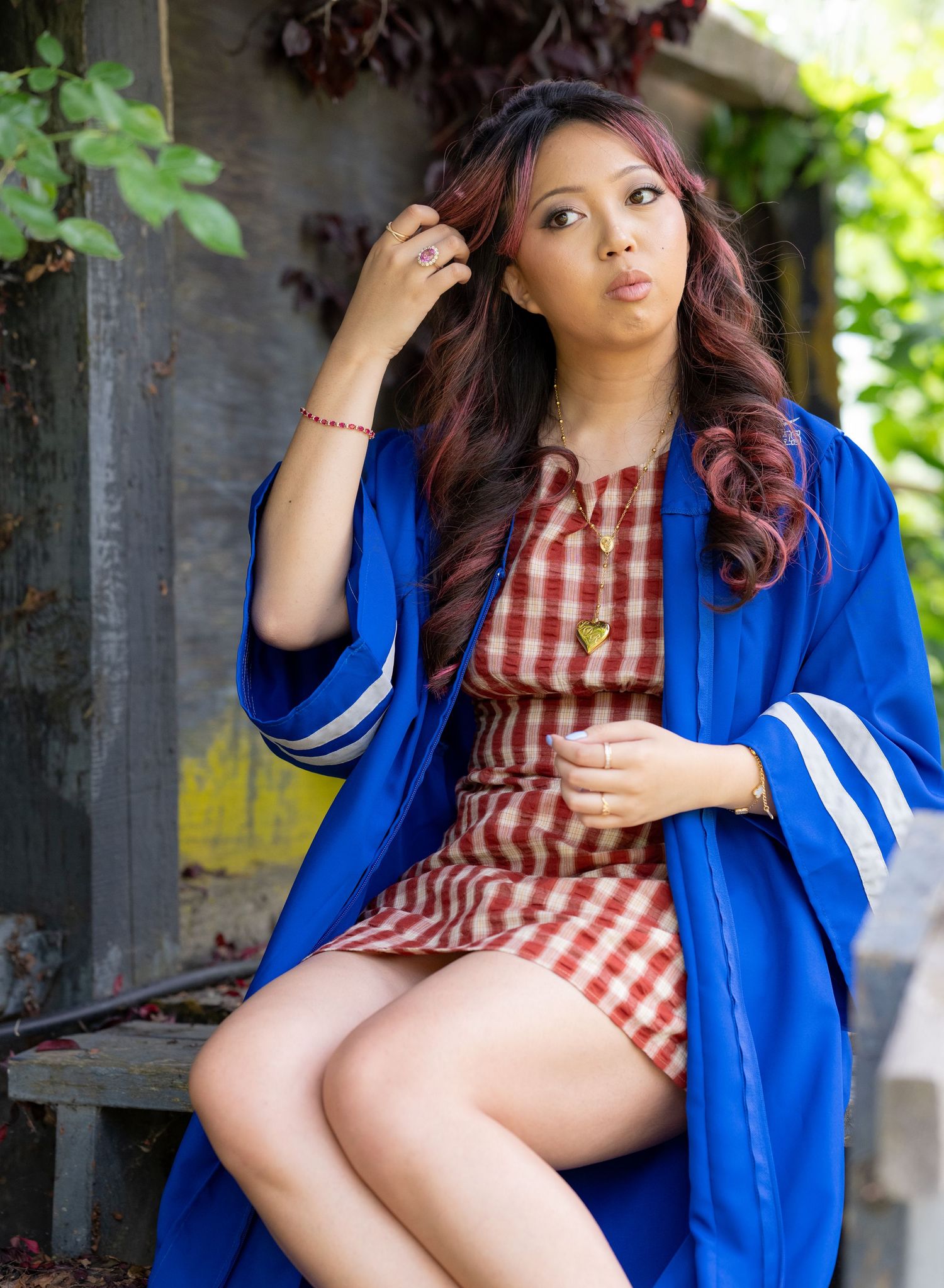 Young woman in blue graduation gown and red gingham dress poses outdoors on bench.