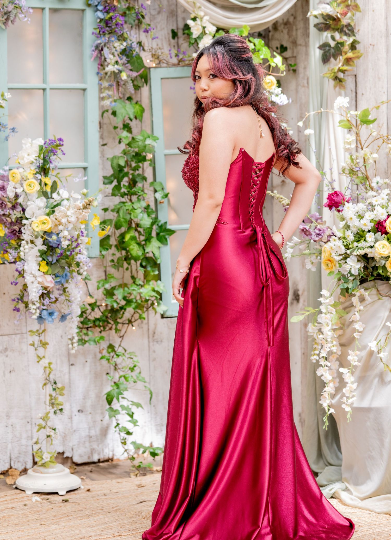 Back view of woman in stunning red satin formal dress with corset lacing and bow detail amid flowers.