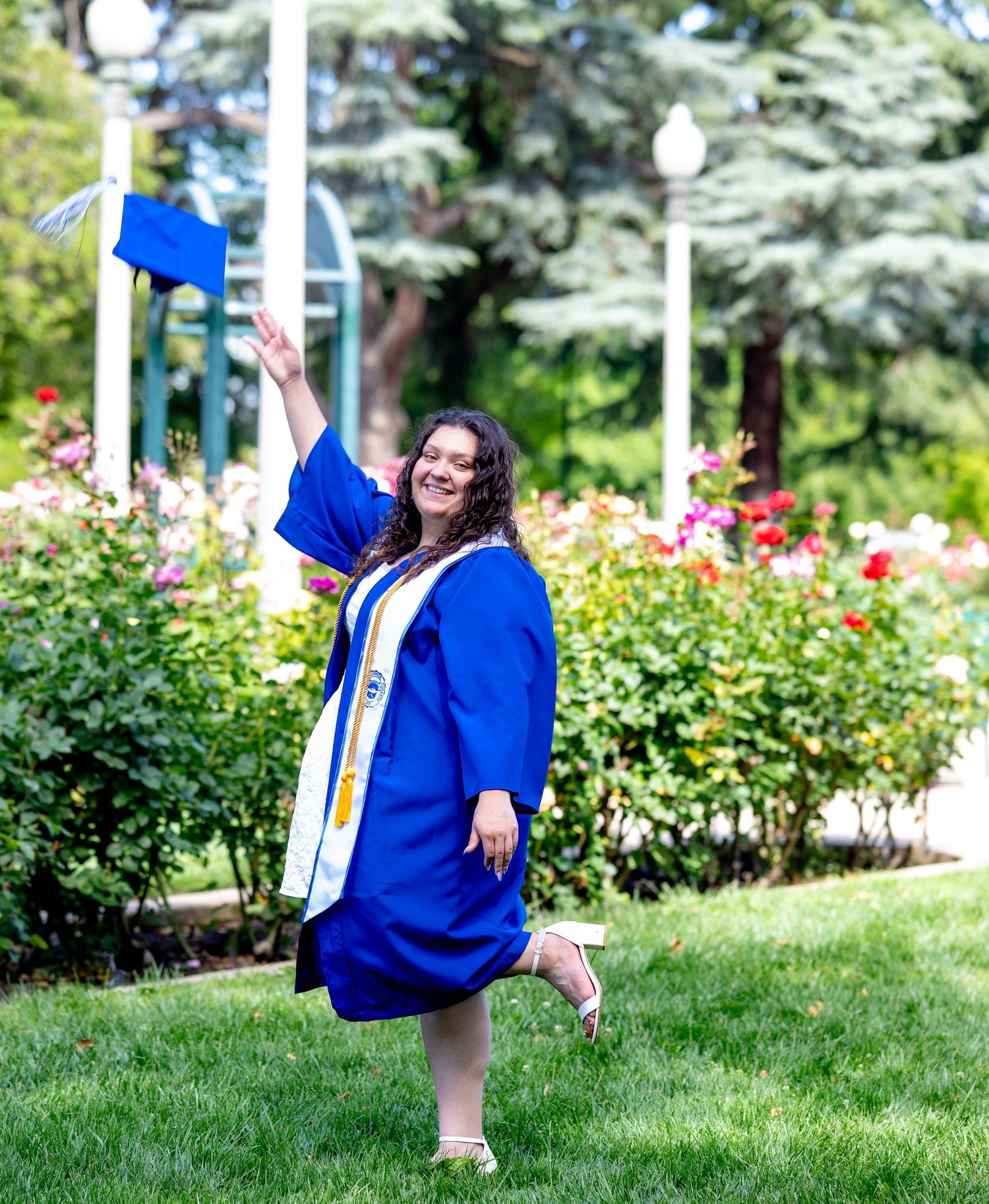 Joyful graduate tosses her blue mortarboard into the air in a vibrant garden.