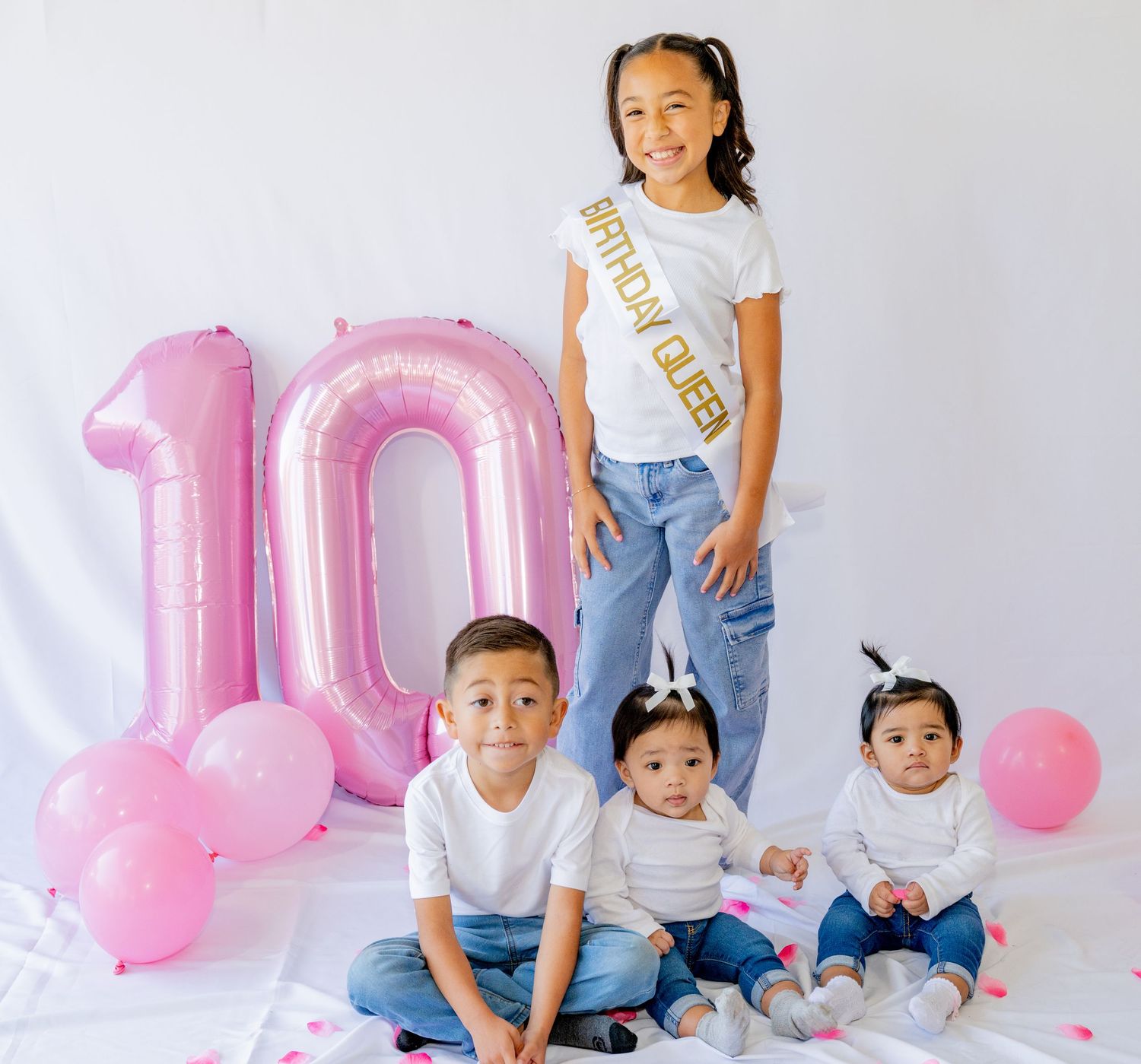 Girl wearing birthday queen sash stands with three younger siblings by pink number 10 balloons.
