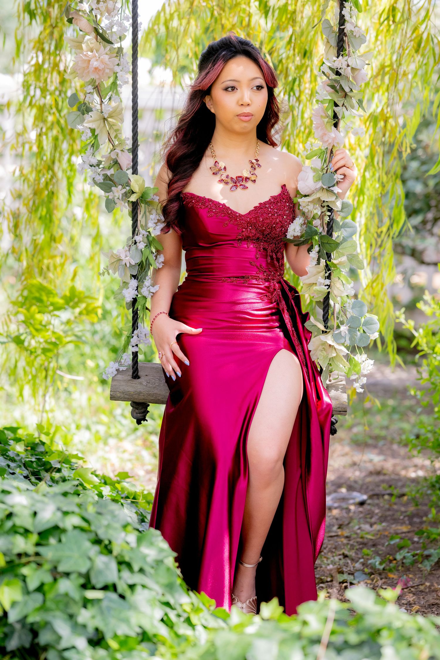 Asian woman in crimson satin gown poses on a flower-adorned swing in green garden.