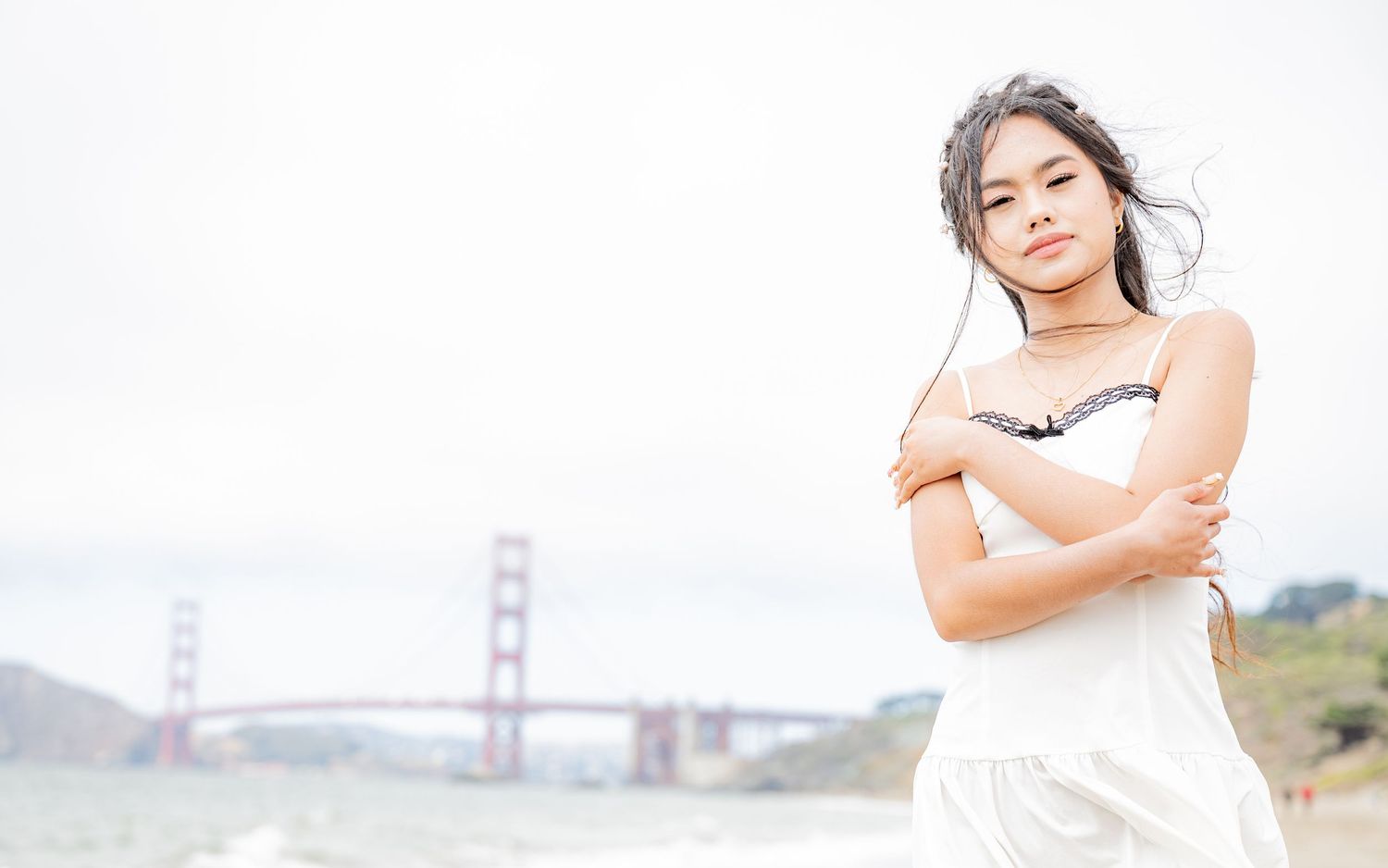 Young woman in white dress with arms crossed poses at Baker Beach near Golden Gate Bridge.