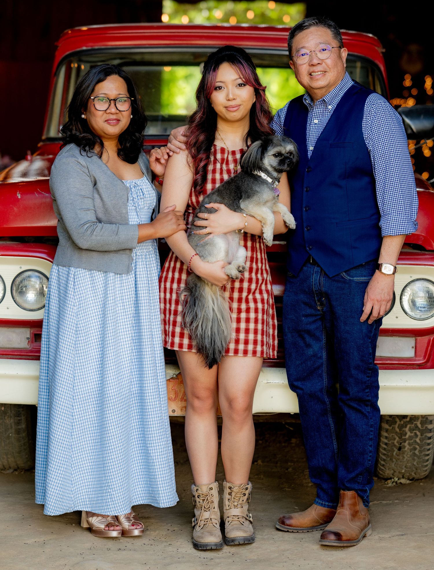 Family portrait in front of vintage red truck, young woman holding gray dog flanked by parents smiling.