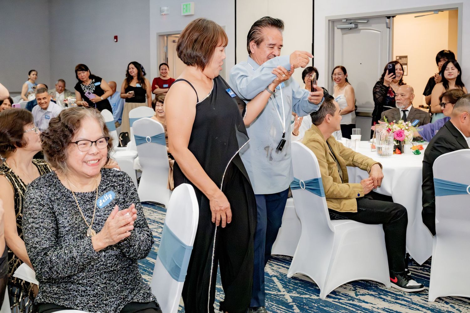 Guests celebrate at an elegant banquet hall event, with a man placing a medal on a woman while attendees applaud.