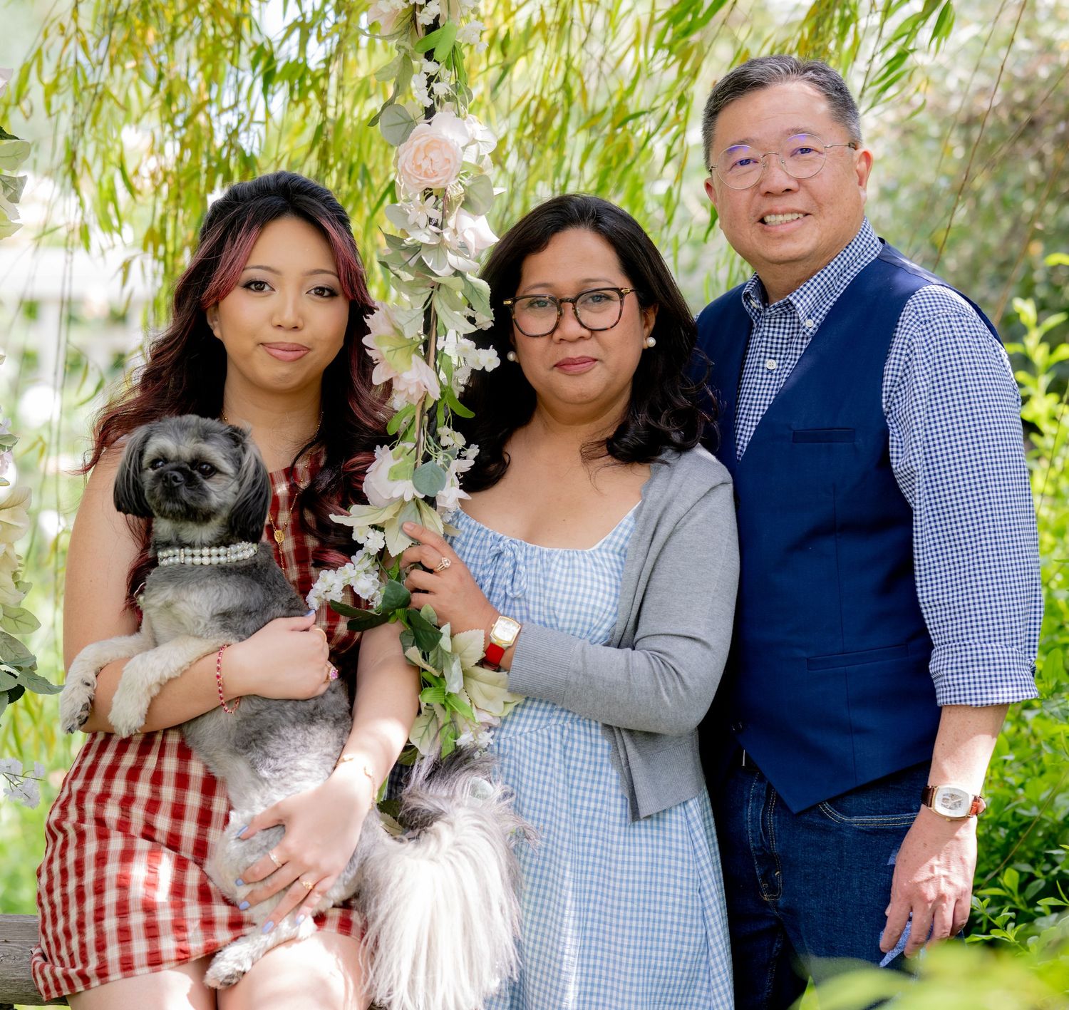 Family of three posing with small gray dog on floral swing under willow tree.