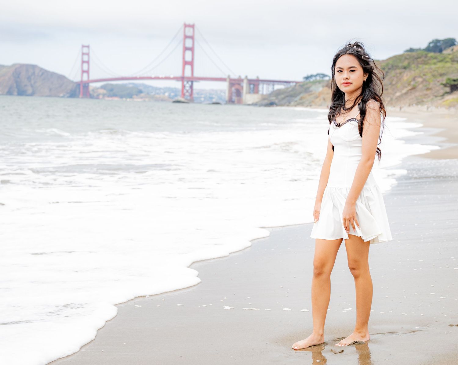 Young woman in white dress standing barefoot on Baker Beach shoreline with Golden Gate Bridge in background.