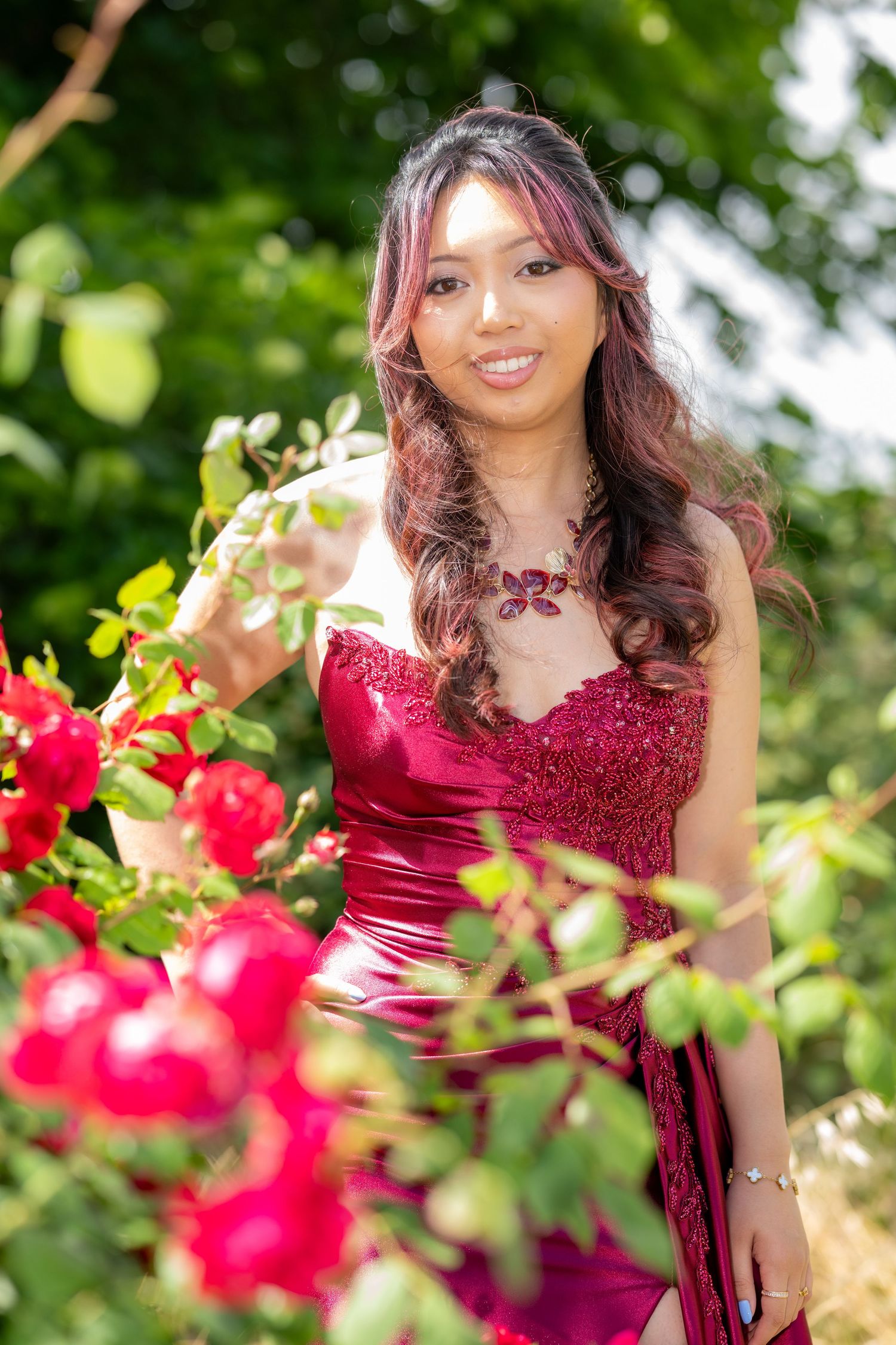 Smiling woman in red satin gown reaches for red roses in sunny garden.