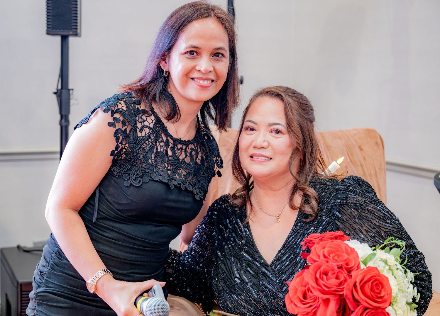 Two women smiling together at a festive birthday party, one holding a red roses bouquet.