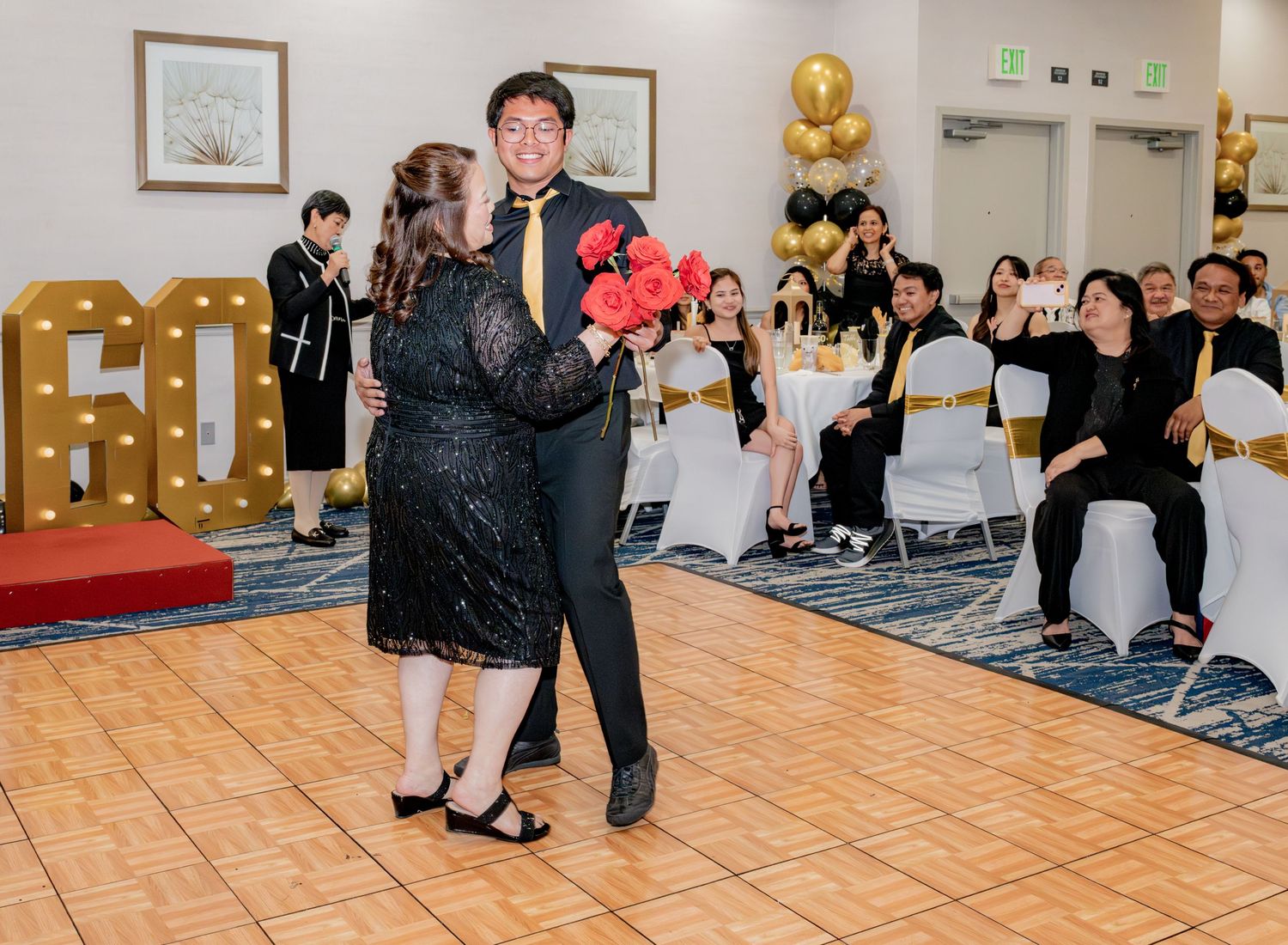 Man in black suit dances with woman holding red roses at elegant 60th birthday party celebration.