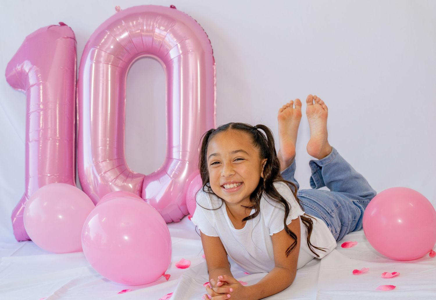 Happy girl lies on white floor smiling beside large pink number 10 birthday balloon.