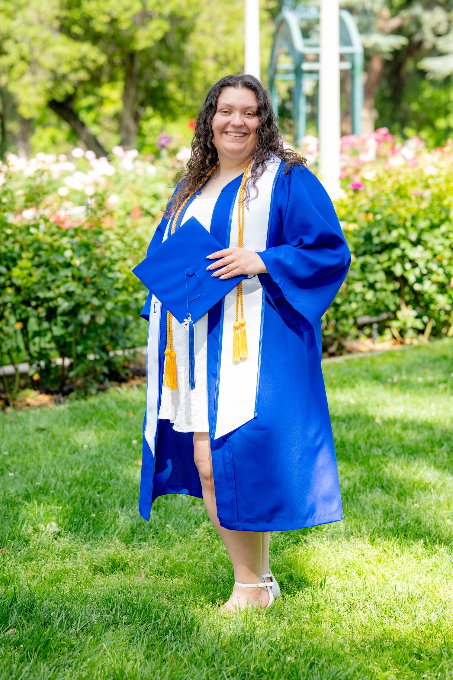 Proud graduate in blue cap and gown holds cap while standing on green grass in a garden.