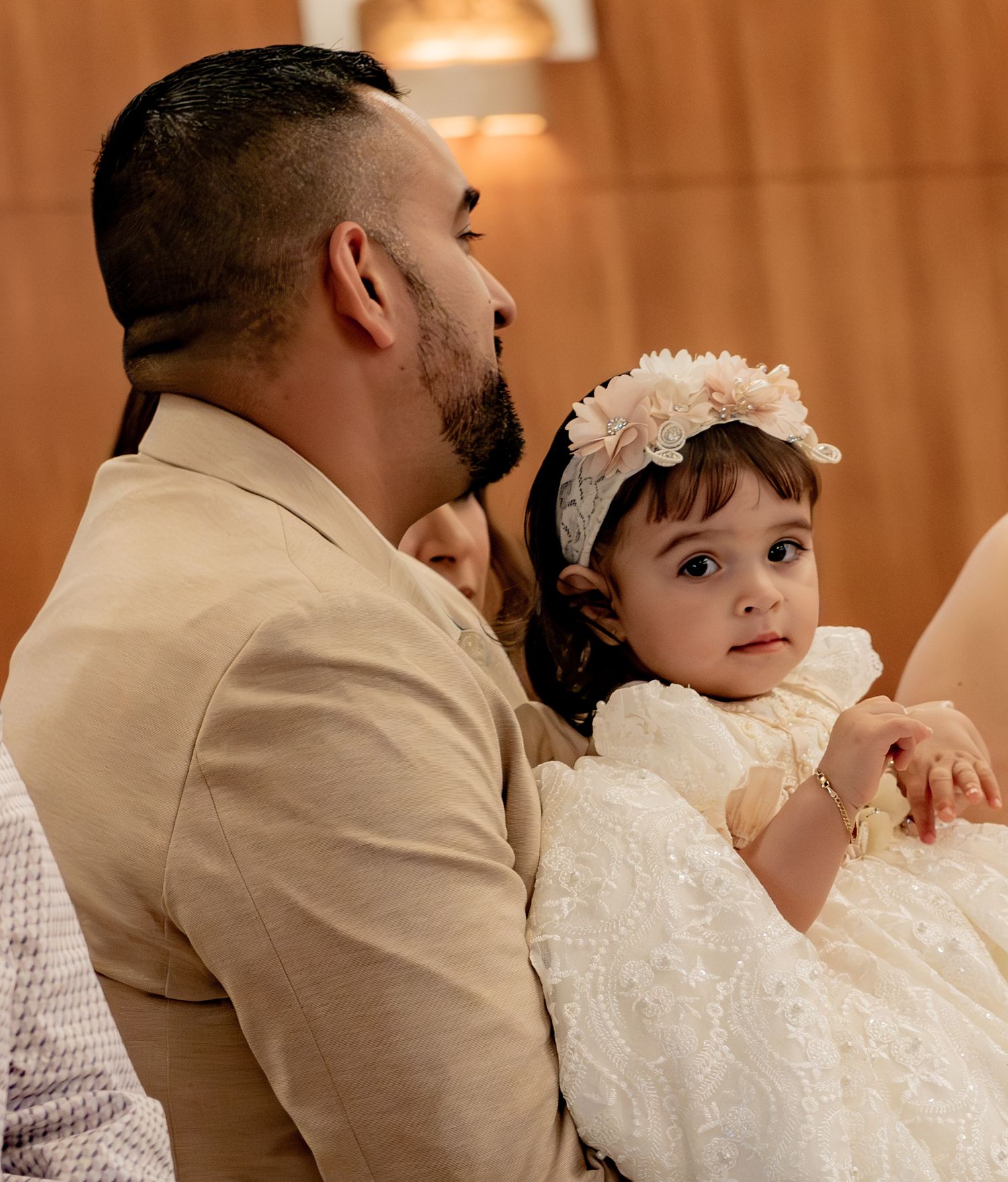 Father holds his baby girl dressed in white lace baptism gown with floral headband in church.