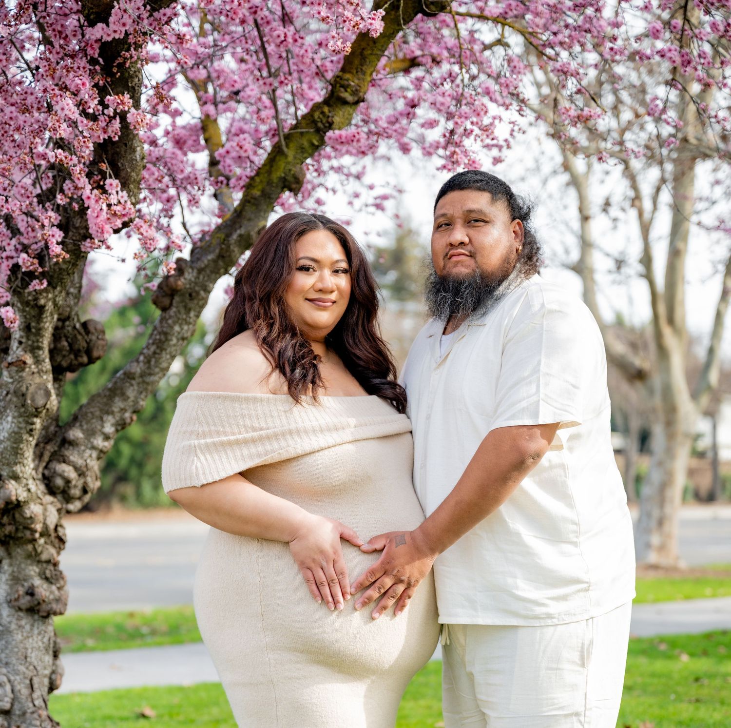 Pregnant couple in cream outfits posing under pink cherry blossom tree on green grass.