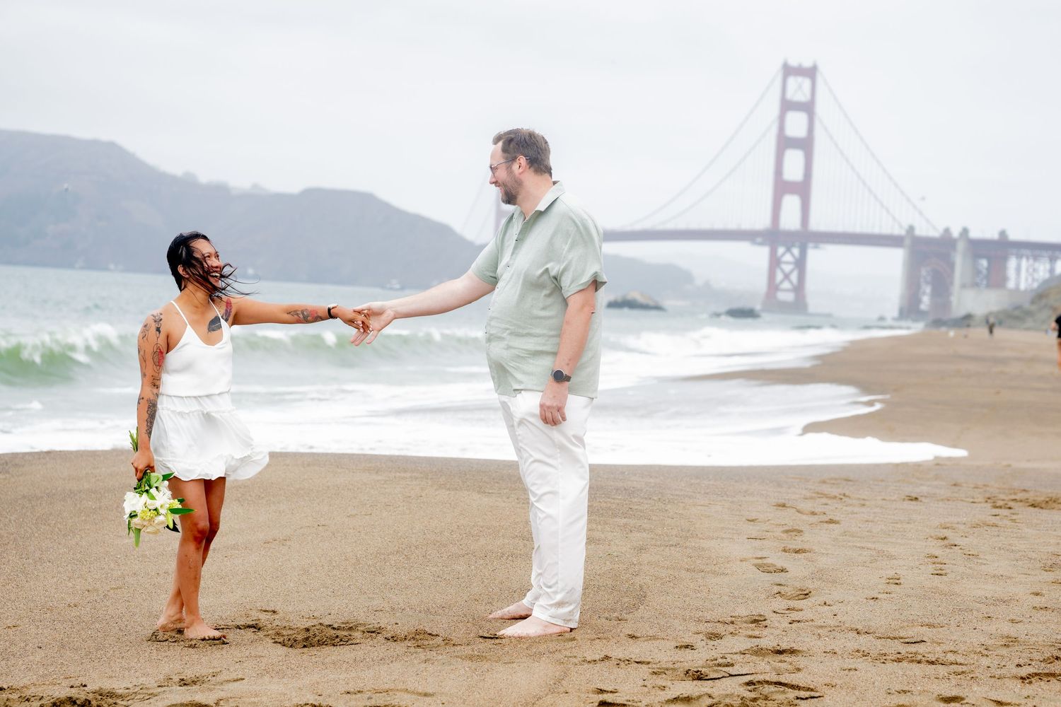 Couple holding hands on Baker Beach with Golden Gate Bridge backdrop, woman holding bouquet.