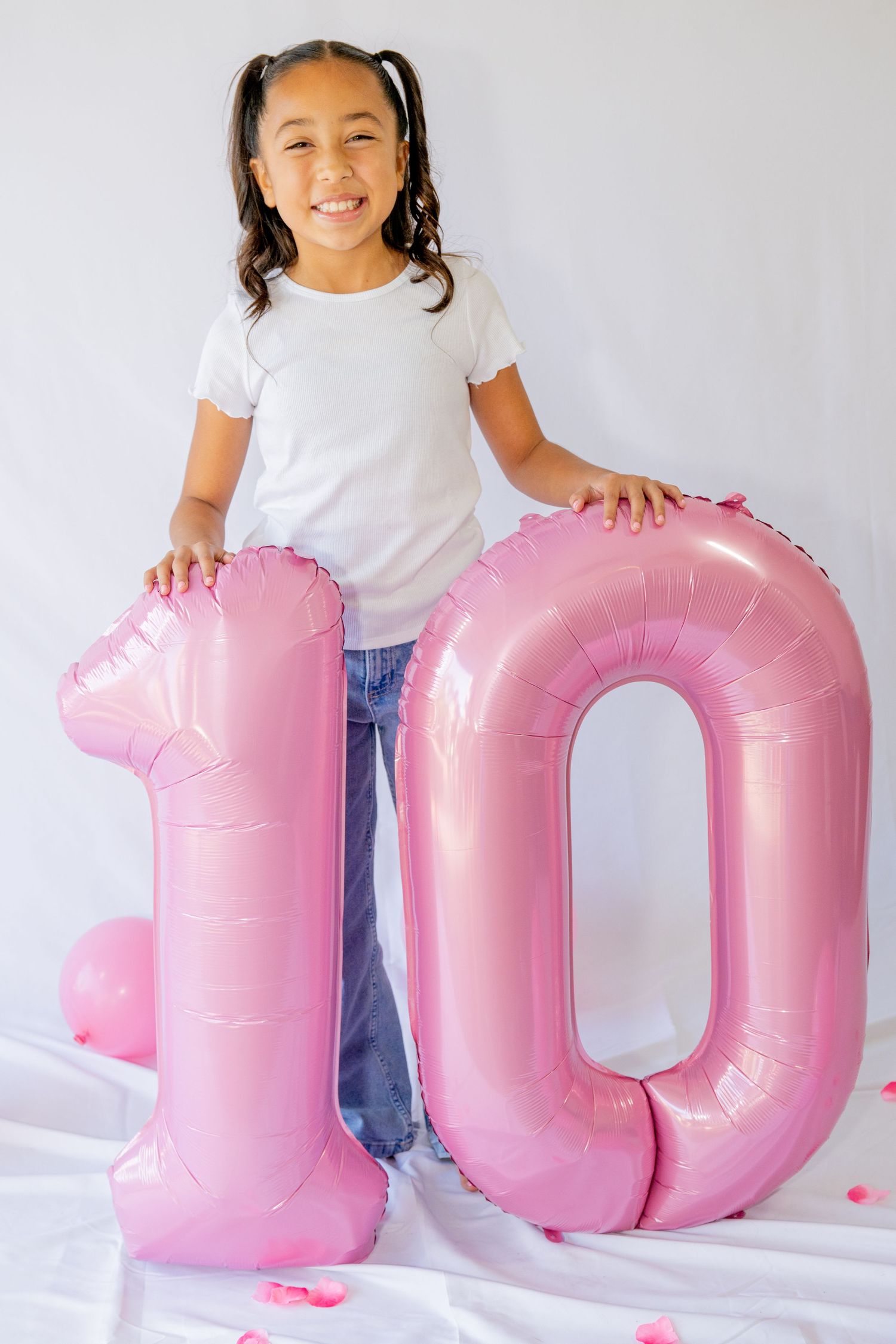 Happy girl standing behind large pink number 10 balloon for birthday celebration on white backdrop.
