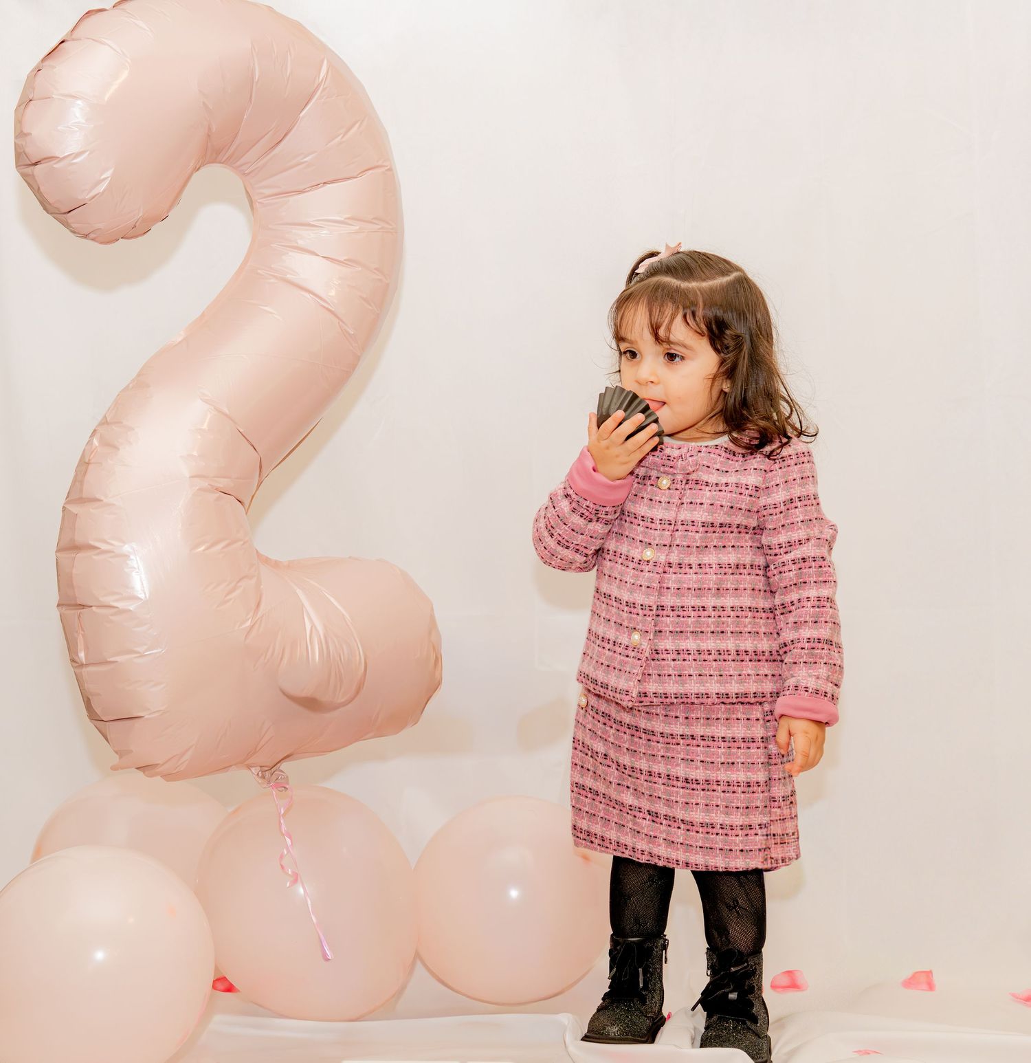 Toddler girl in pink tweed dress eating cupcake beside large number 2 balloon at 2nd birthday party.