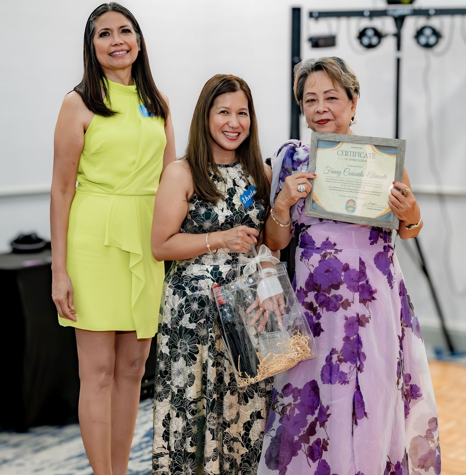 Three women posing together at an awards ceremony, one holding a certificate and gift.